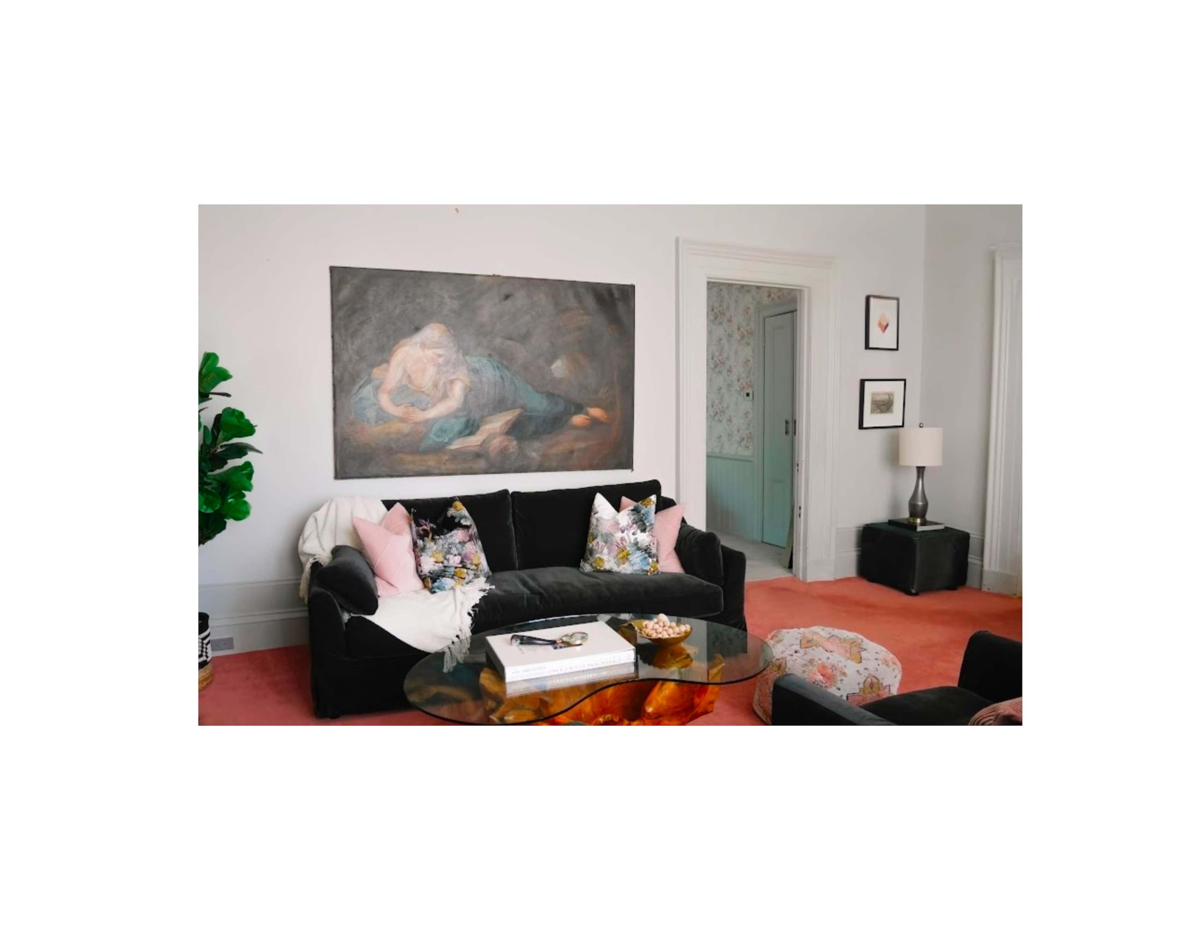 A black sofa with patterned pillows sits in a living room featuring a large painting above, a coffee table in front, and a doorway leading to another room in the background.