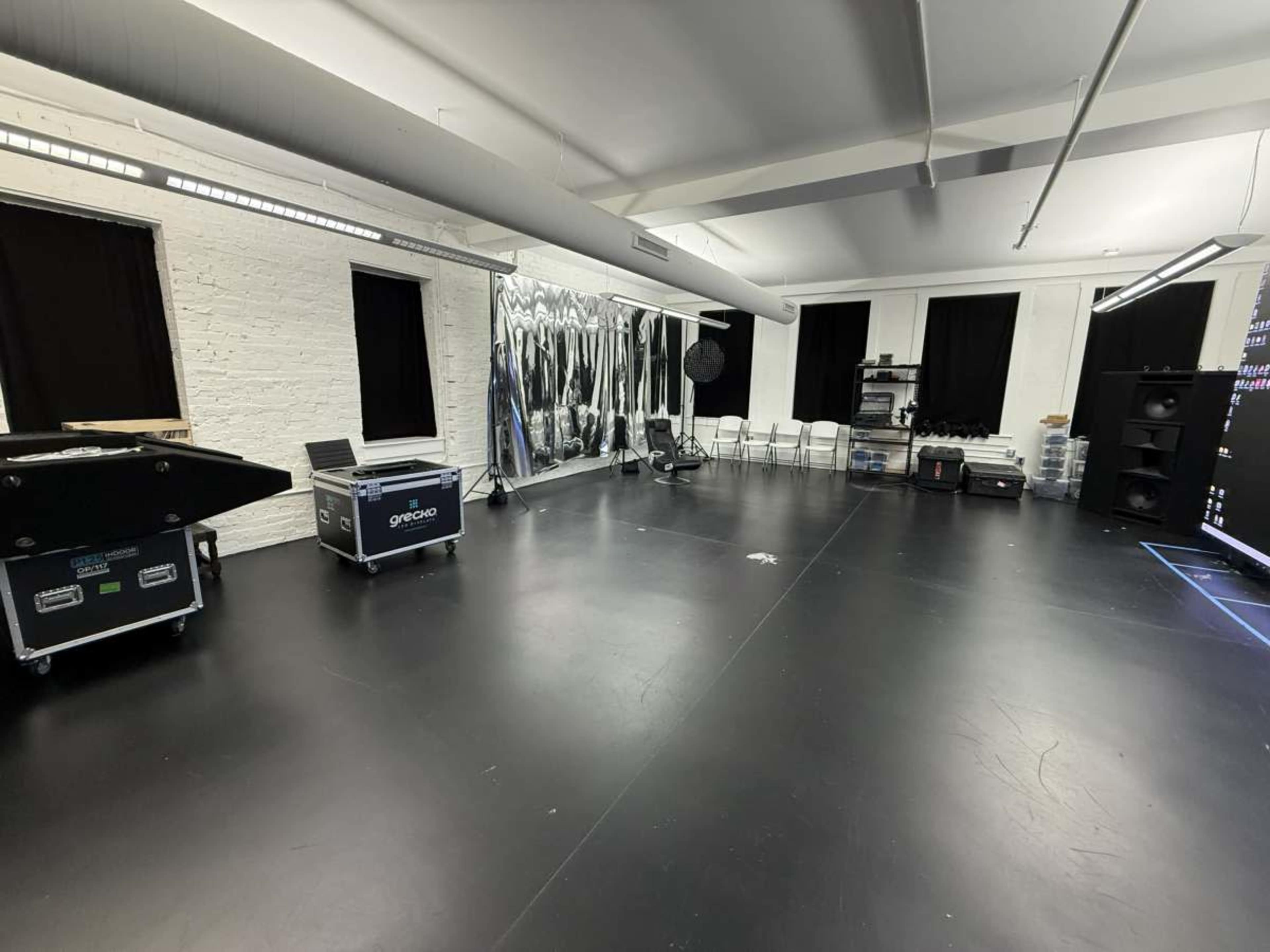 The image shows an empty studio space with a black floor, white brick walls, and various equipment and seating arranged around the room.