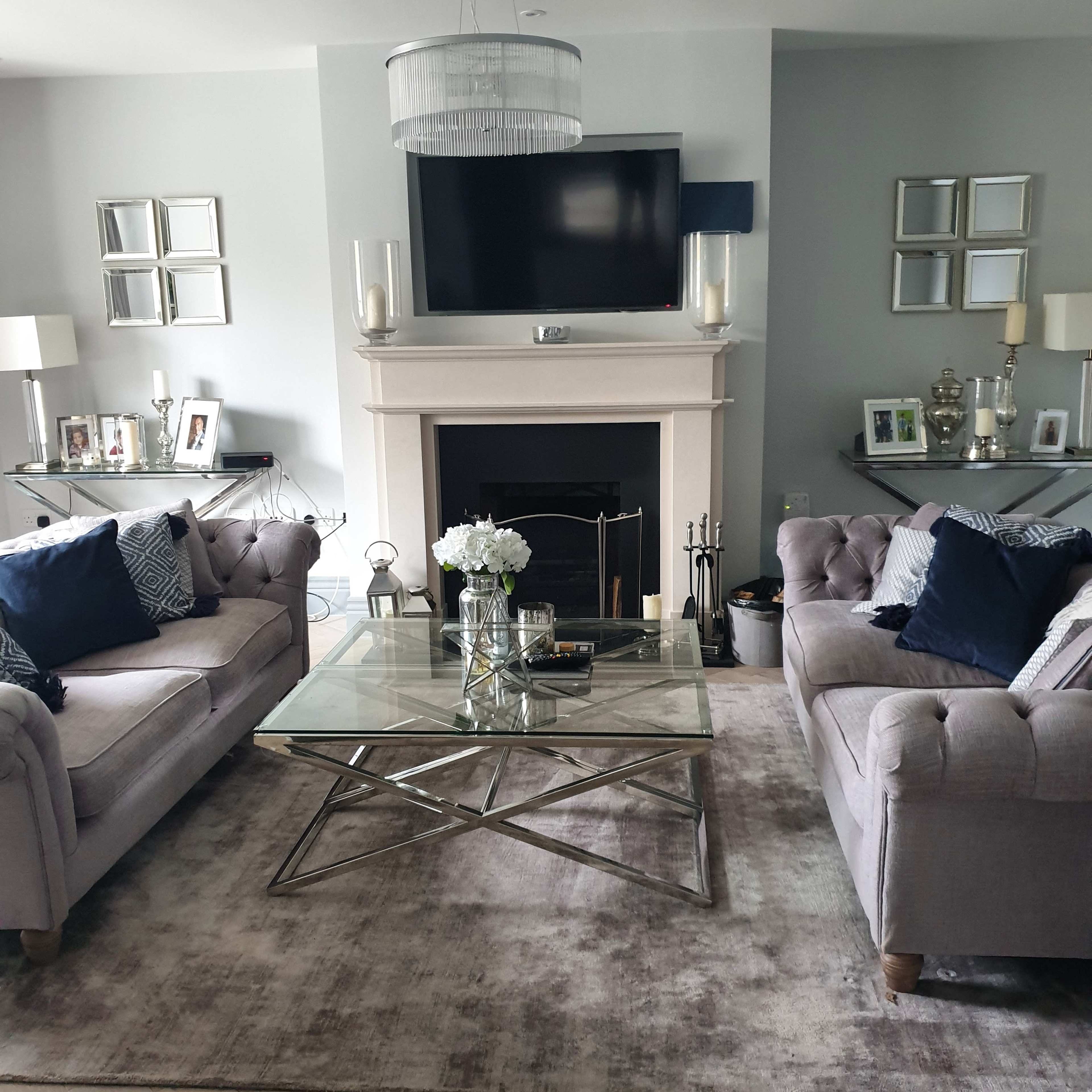 The image shows a living room with two gray tufted sofas, a glass coffee table, and a fireplace with a mounted television above it.