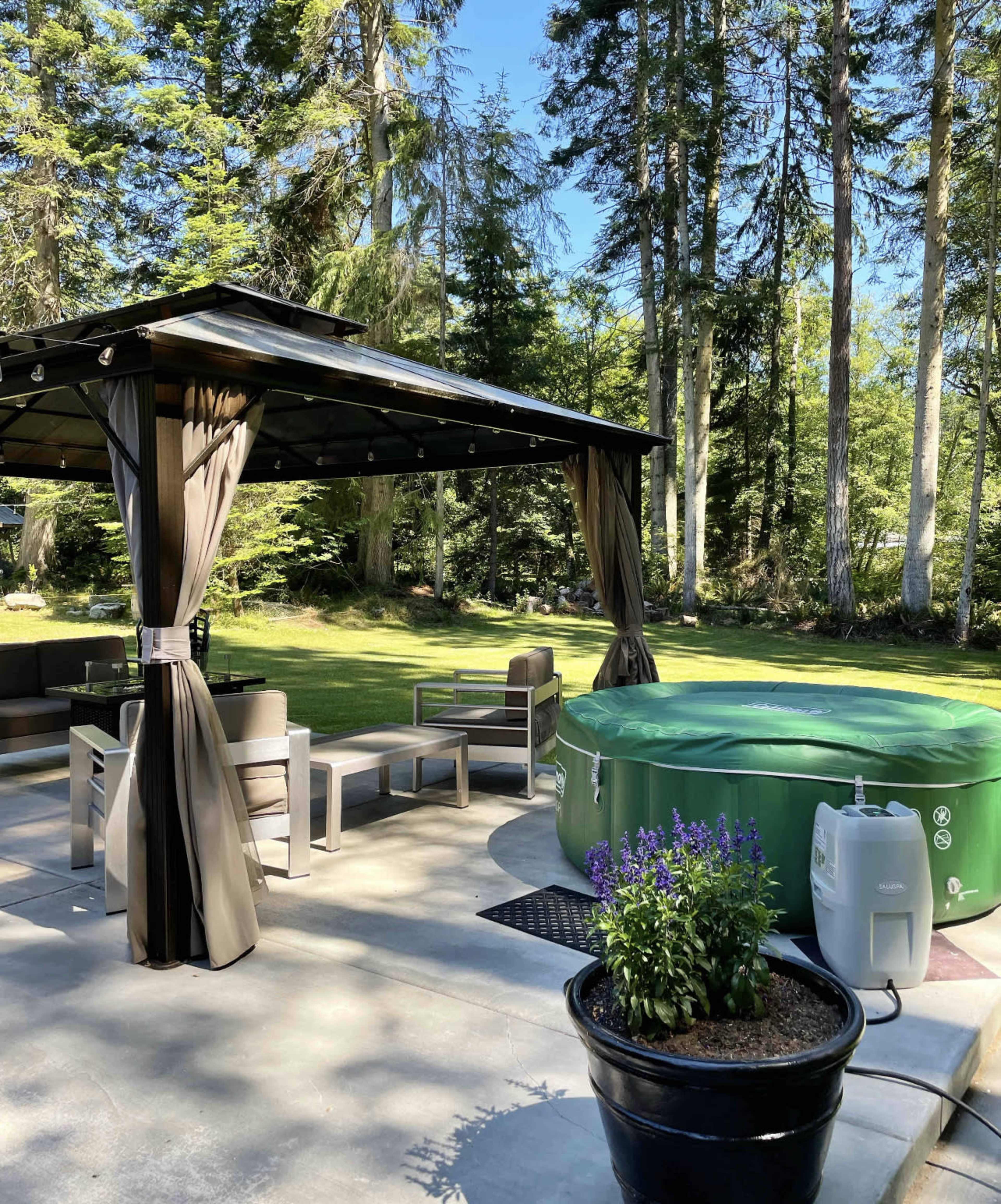 A gazebo with seating surrounds a green hot tub in a forested outdoor area.