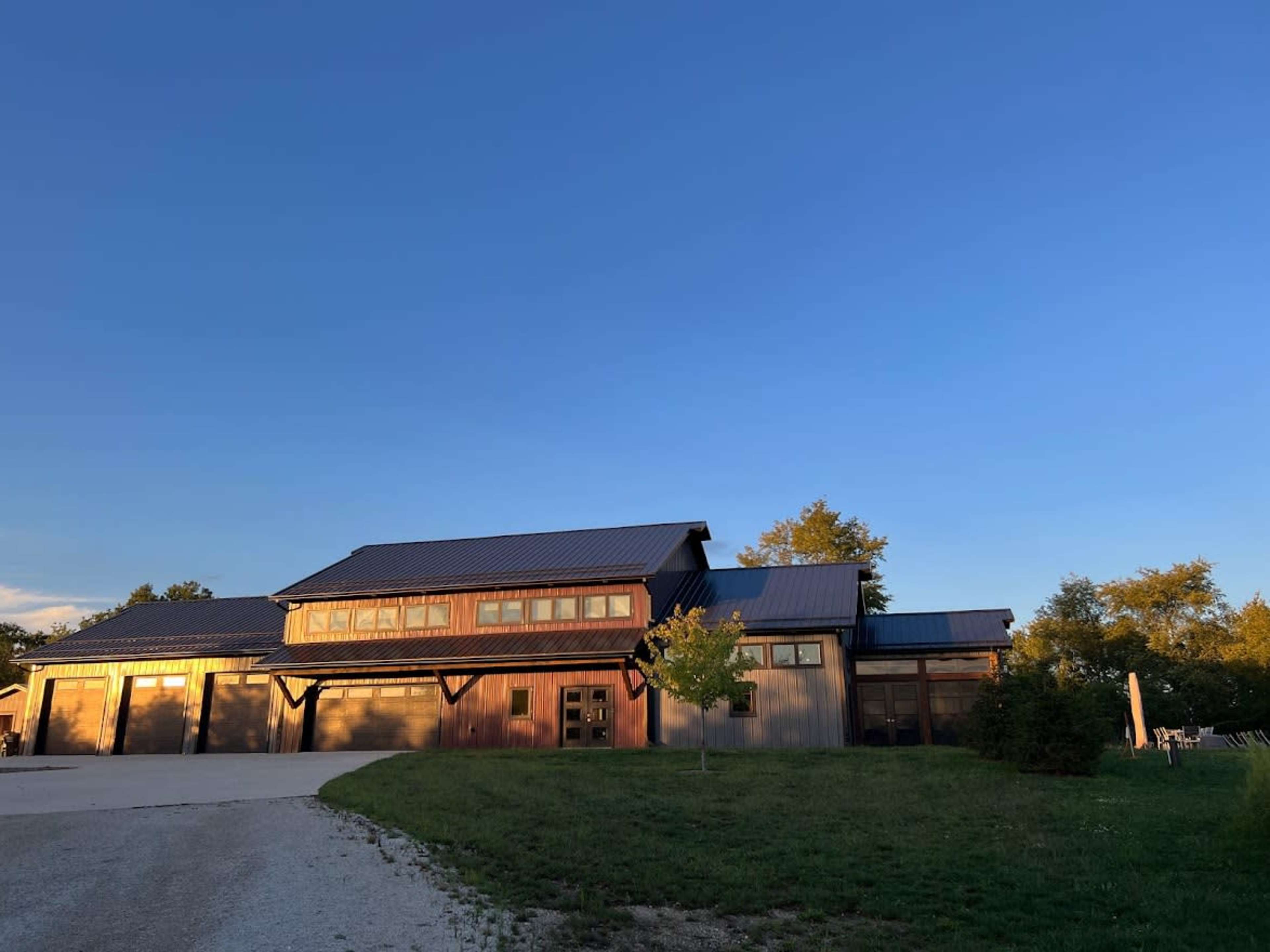 A modern wooden house with large windows and a metal roof is situated on a grassy area under a clear blue sky.
