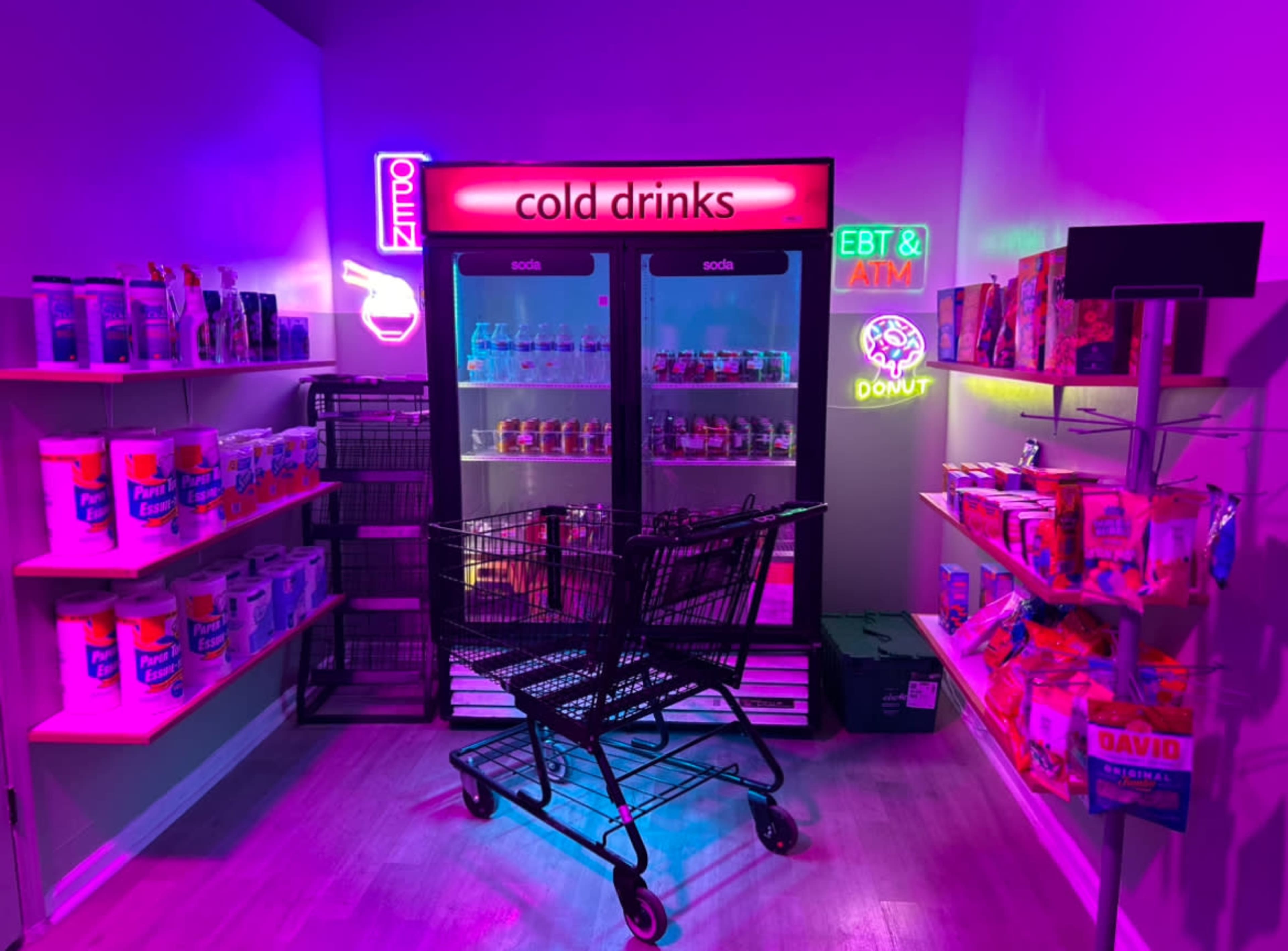The image shows a brightly lit convenience store corner with shelves stocked with snacks and a drinks cooler, along with a shopping cart in the aisle.