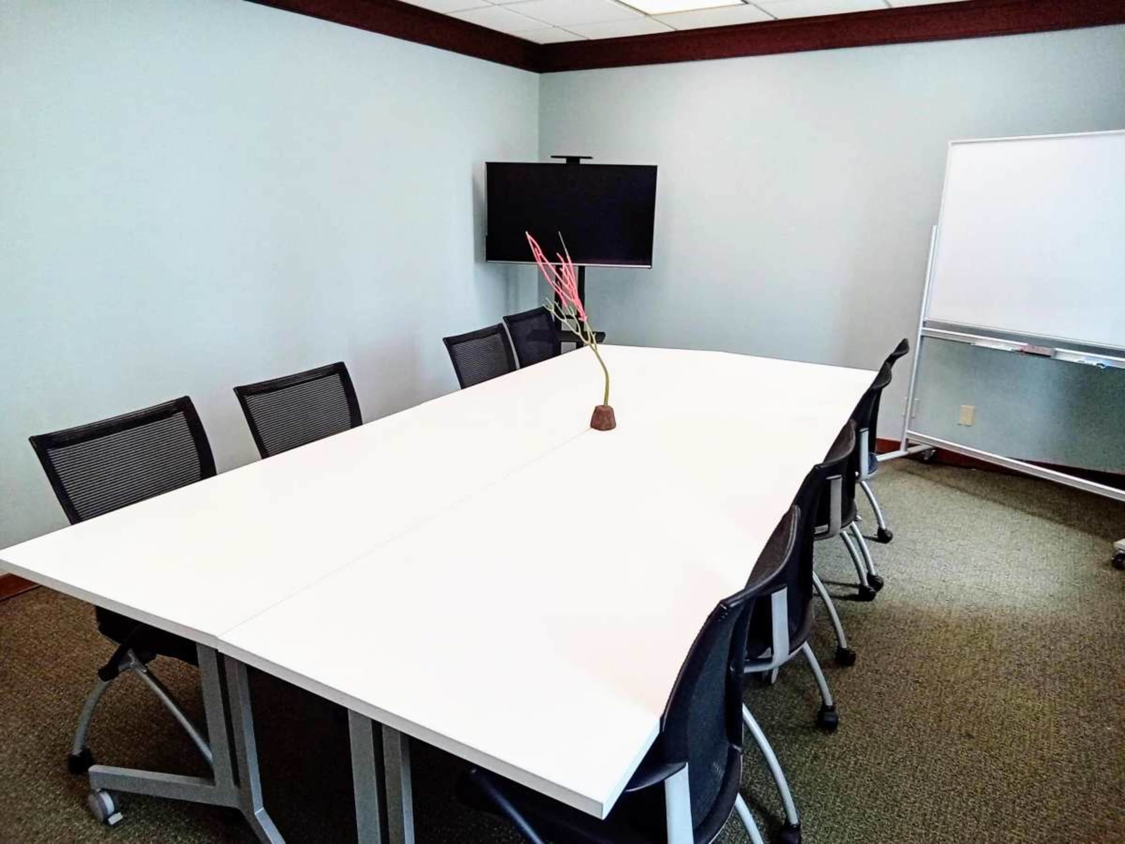 A conference room features a large white table surrounded by black chairs, with a television and a whiteboard against the walls.