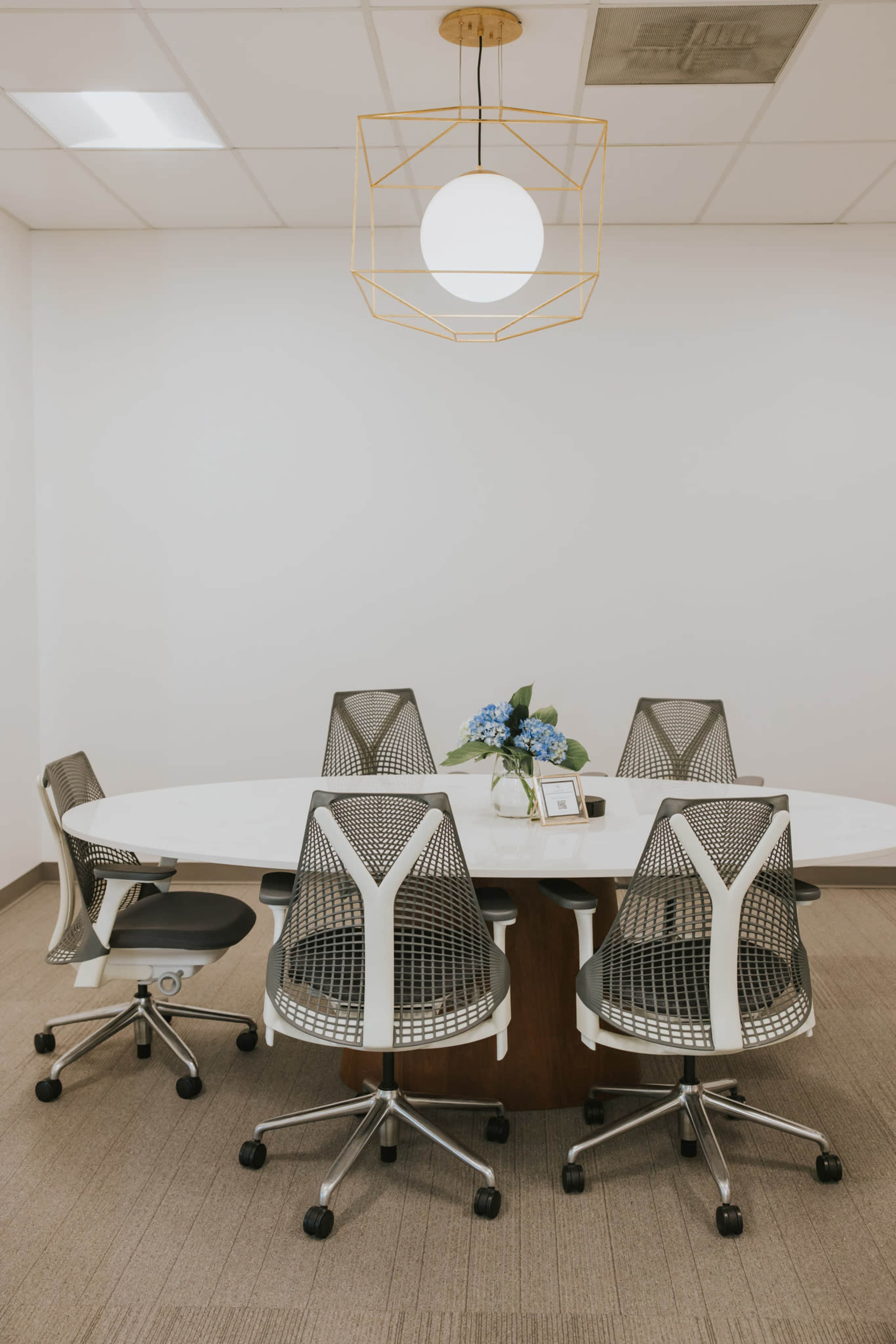 The image shows a modern conference room with a large oval table surrounded by six ergonomic chairs, illuminated by a pendant light.