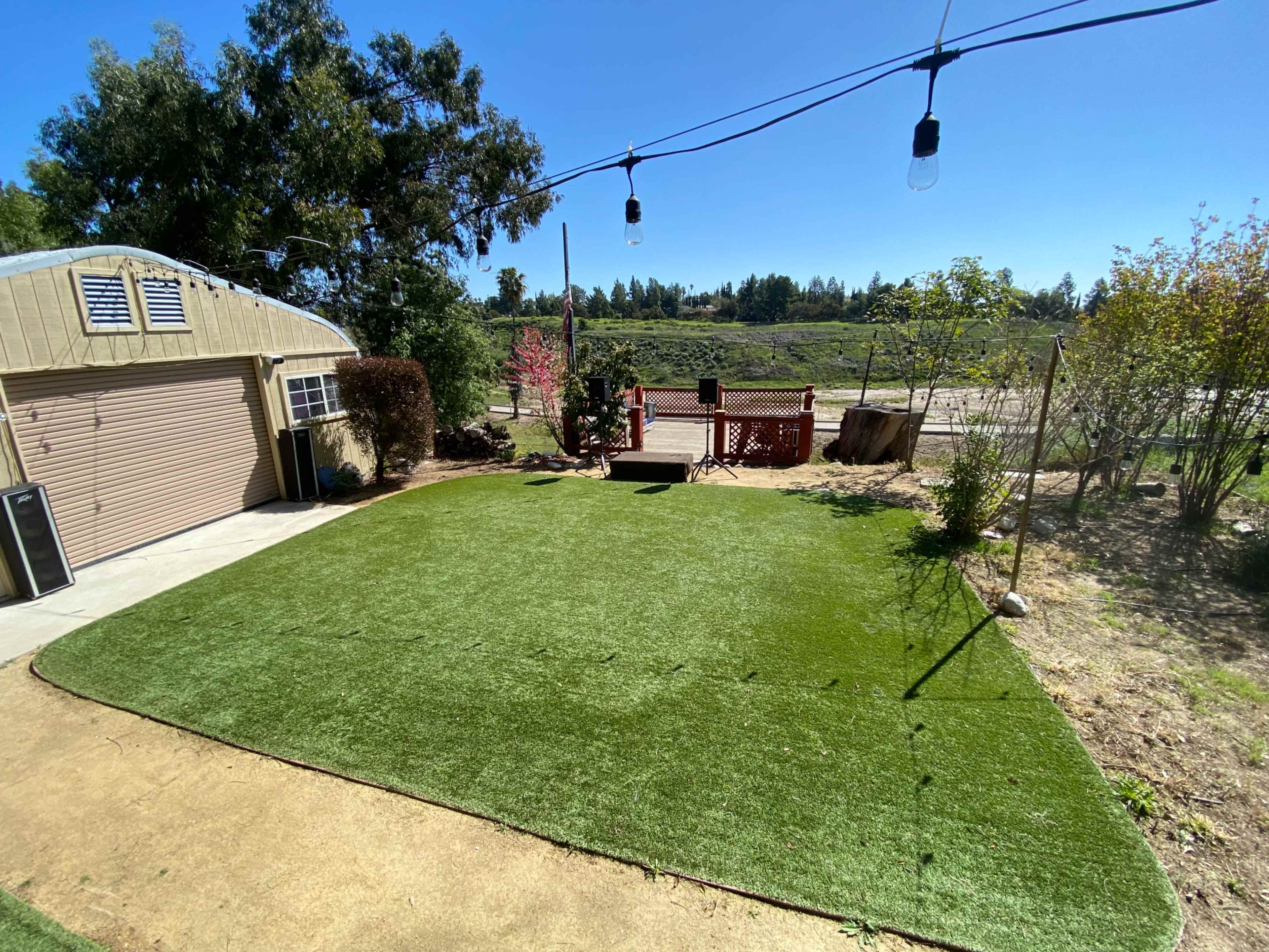 A backyard with artificial grass, a small wooden deck, and a garage, bordered by a pathway and trees in the background.