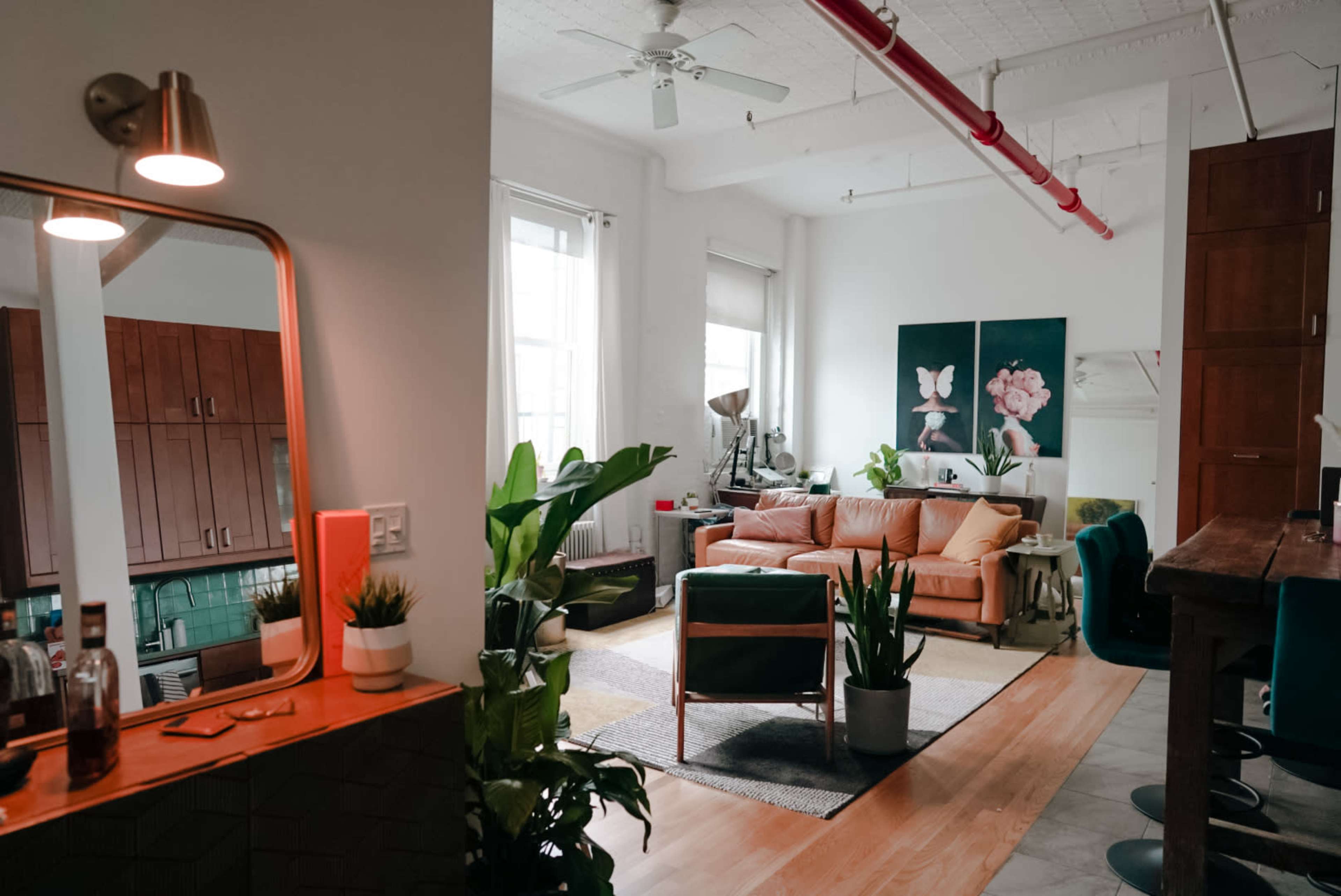A cozy living room features a peach-colored sofa, large plants, and artwork on the walls, with a view of a kitchen area in the background.