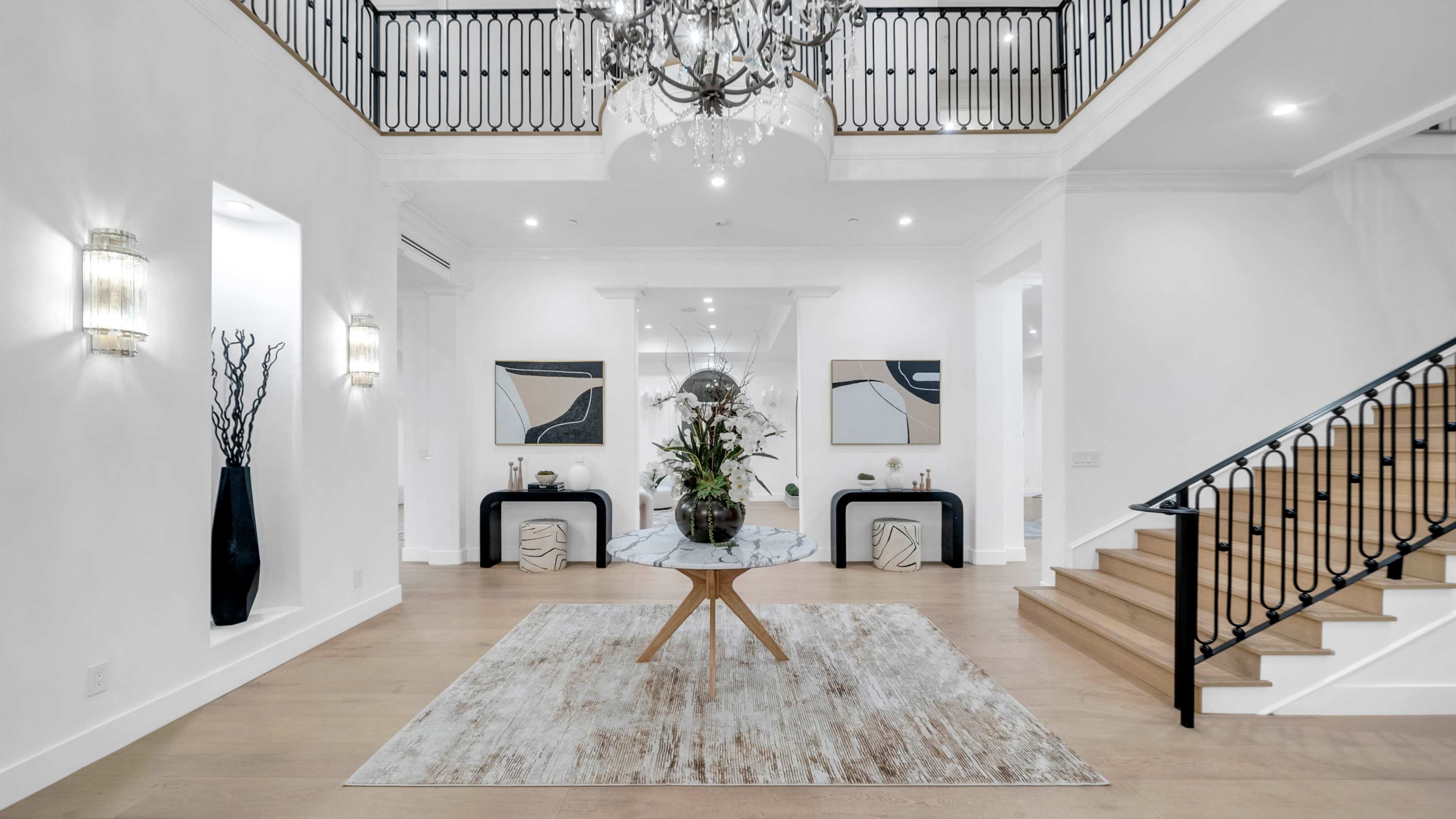 The image depicts a spacious foyer featuring a central round table adorned with floral arrangements, two abstract paintings on the walls, and a staircase leading to an upper level.