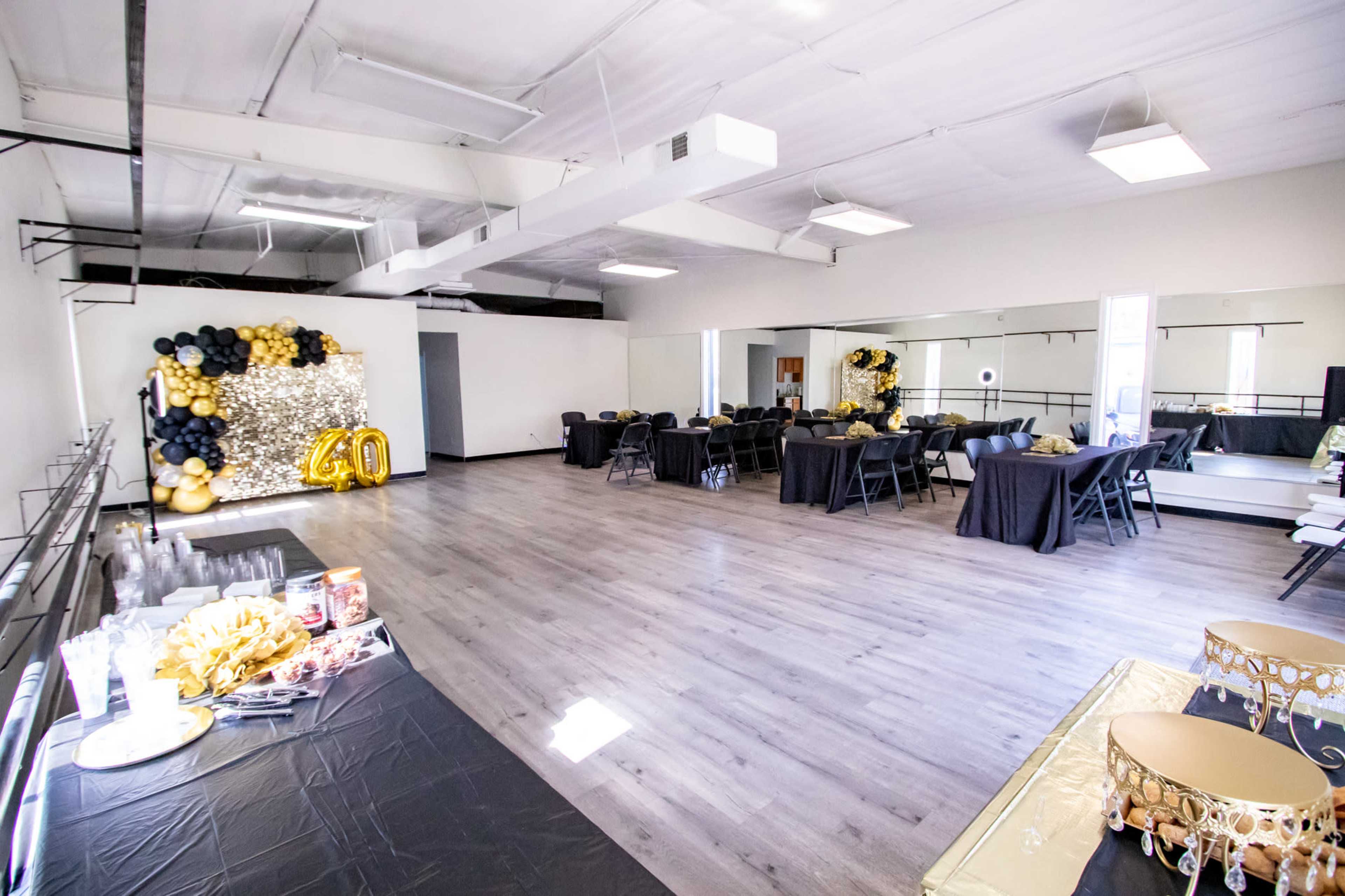 A spacious event room is set up with black tables, gold decorations, and a backdrop of balloons for a celebration.