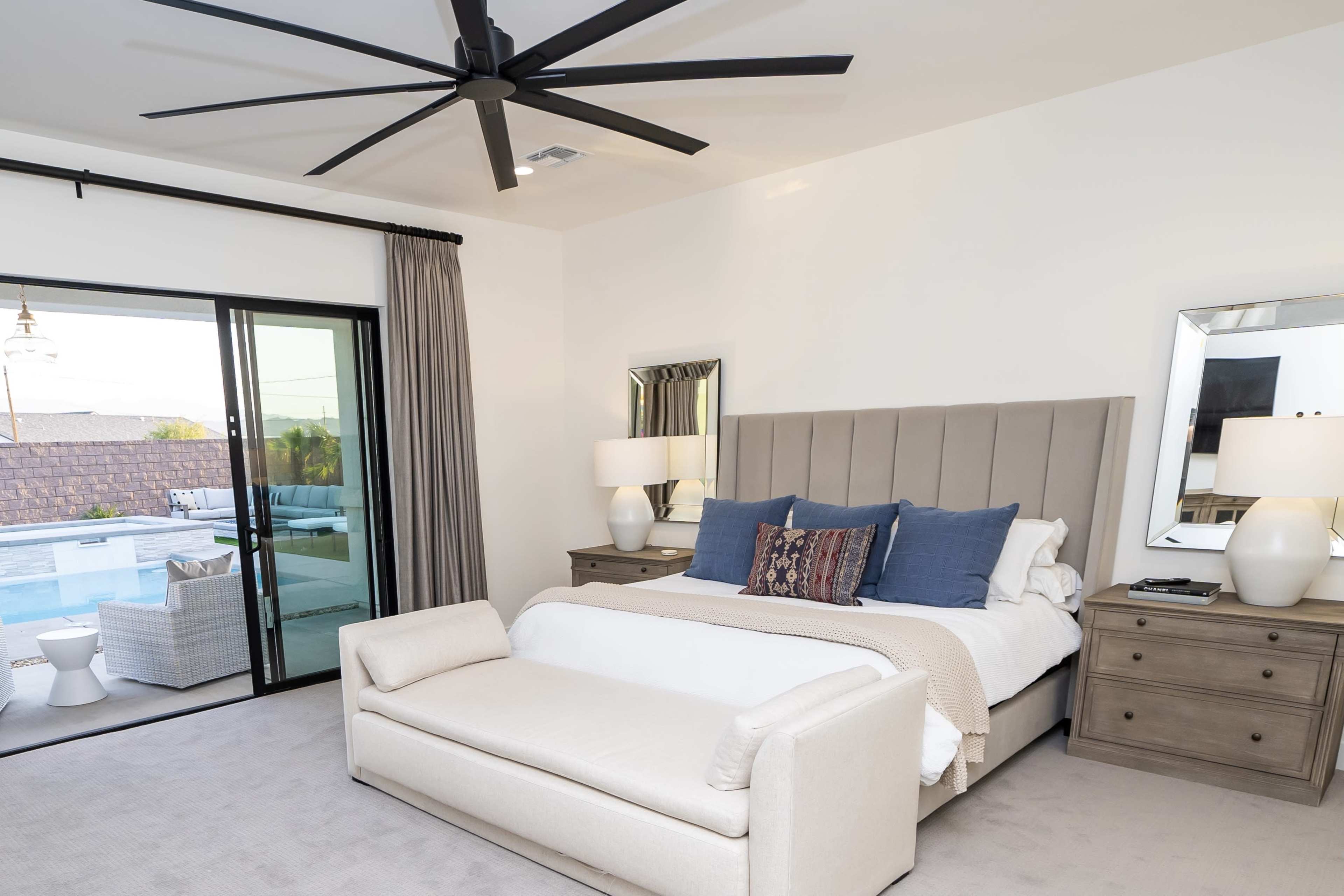 A modern bedroom featuring a large bed with a tufted headboard, a seating area at the foot of the bed, and sliding glass doors leading to an outdoor space with a pool.
