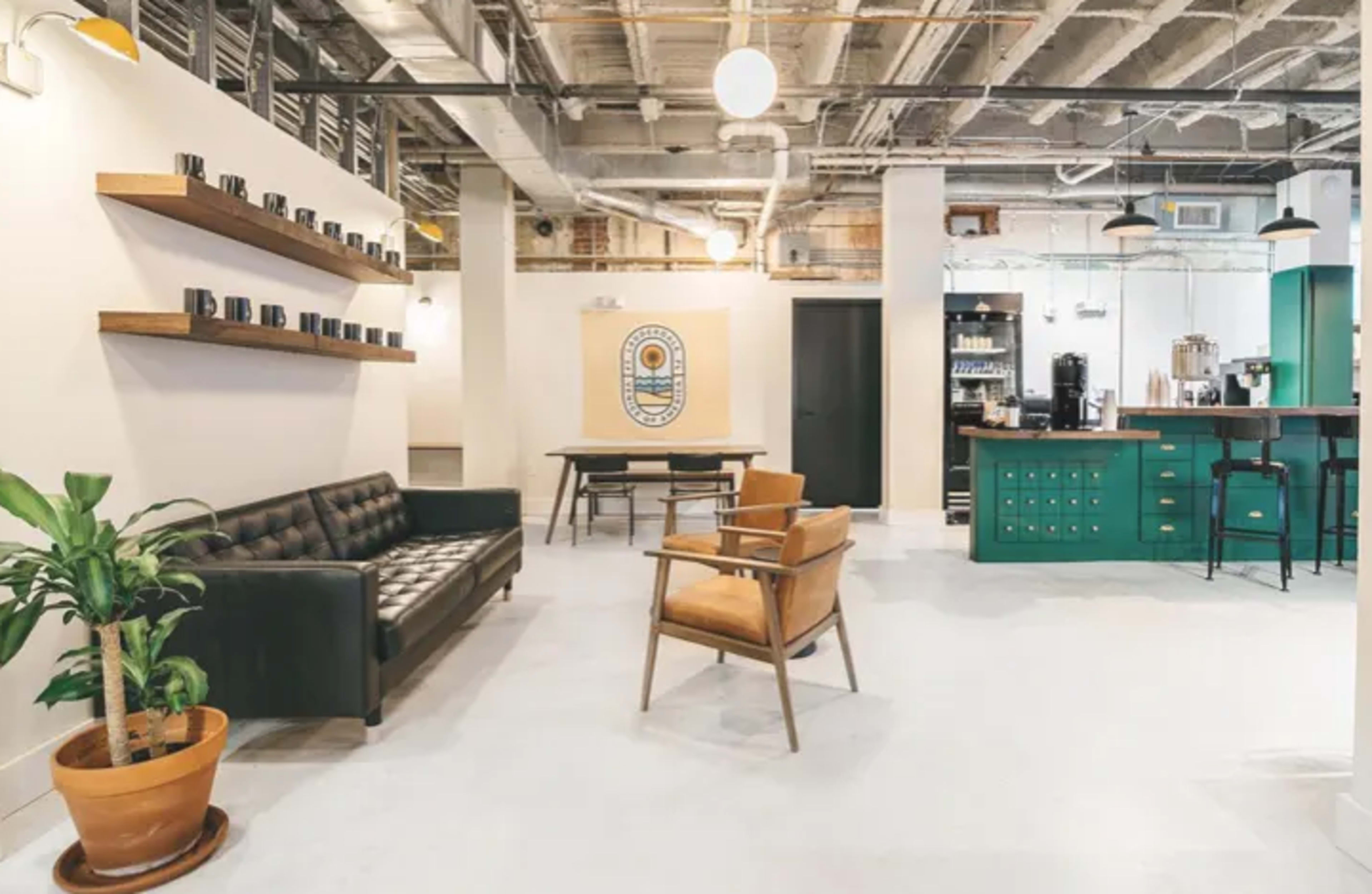 The image shows a modern office space featuring a black couch, brown chairs, a shelf with mugs, and a kitchenette with green cabinetry.