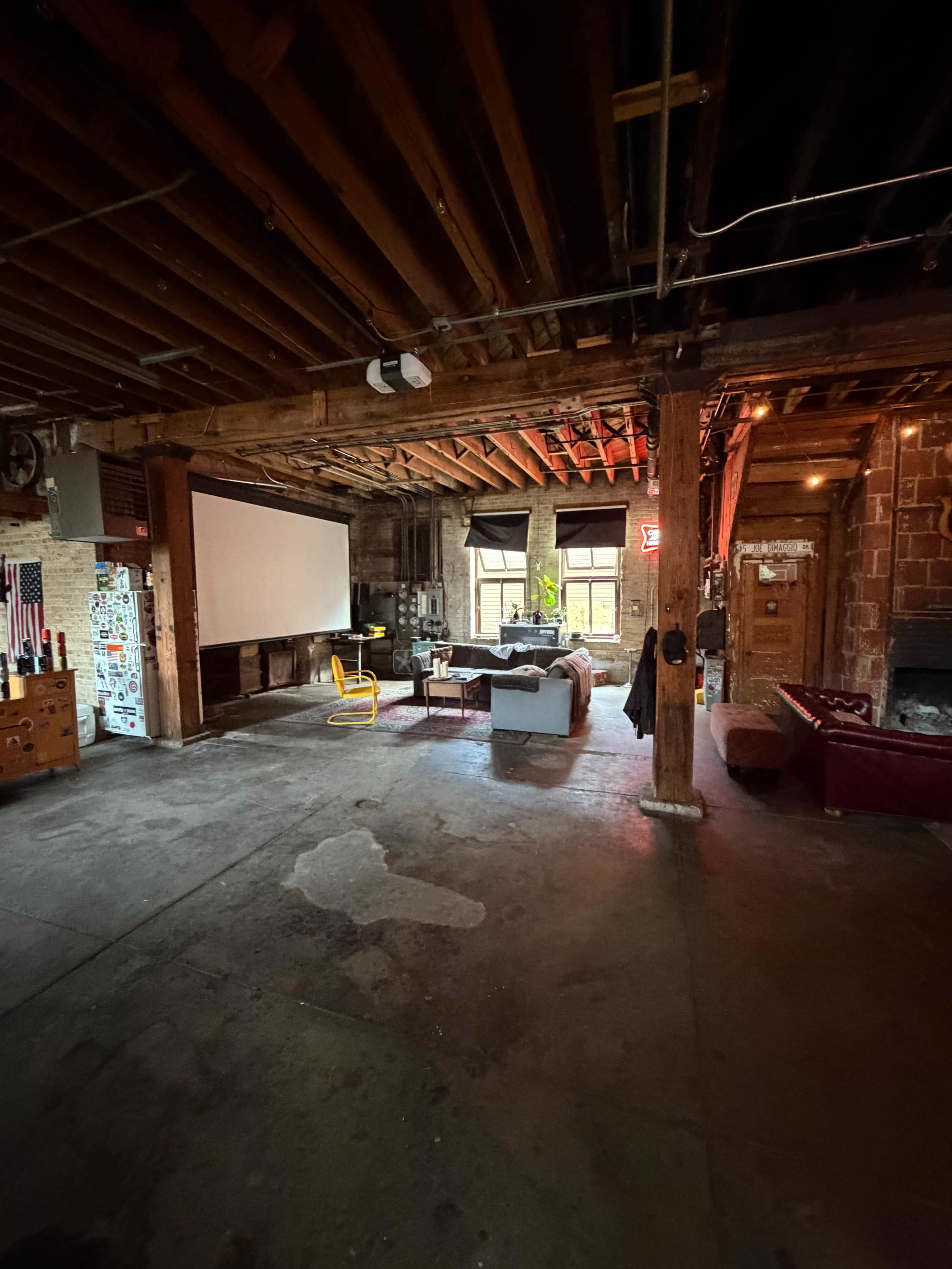 The image shows a spacious, industrial-style room with exposed wooden beams, a projector screen, a sofa, a yellow chair, and scattered furniture.