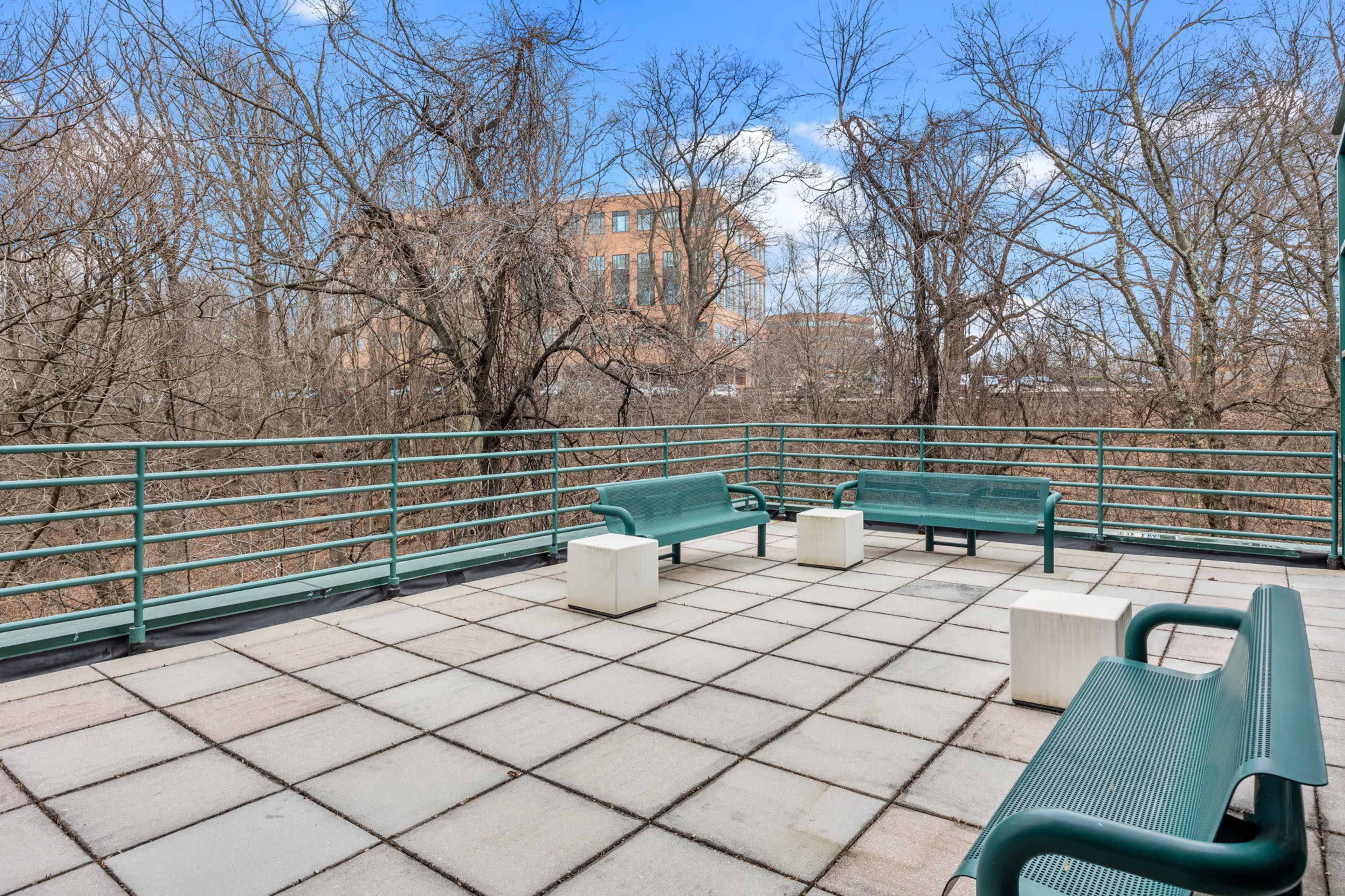 A rooftop terrace features several green benches and concrete tables surrounded by bare trees and a building in the background.