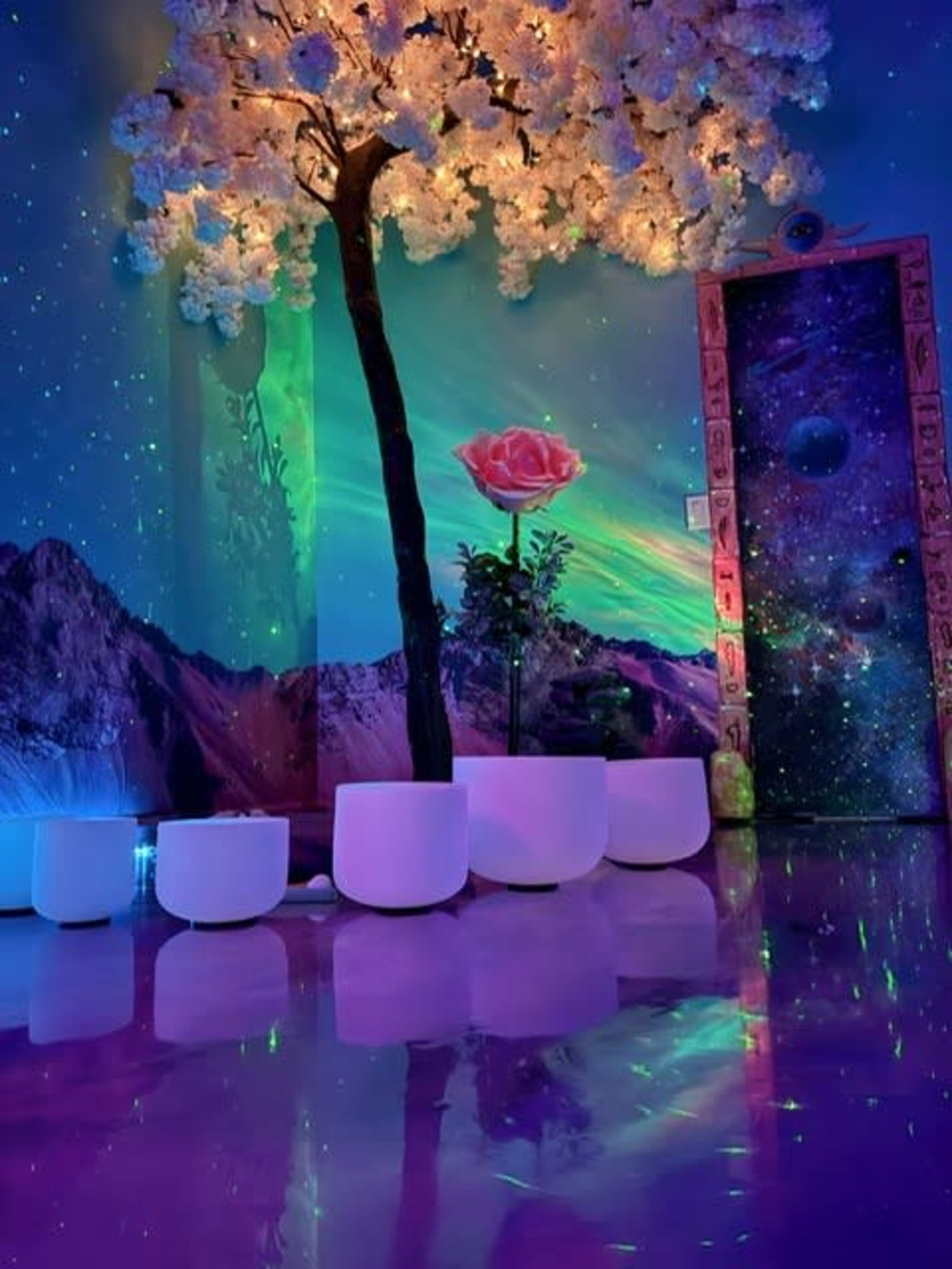 A colorful, whimsical room featuring a large illuminated tree, a rose in a vase, and a mural of mountains and cosmic scenery.