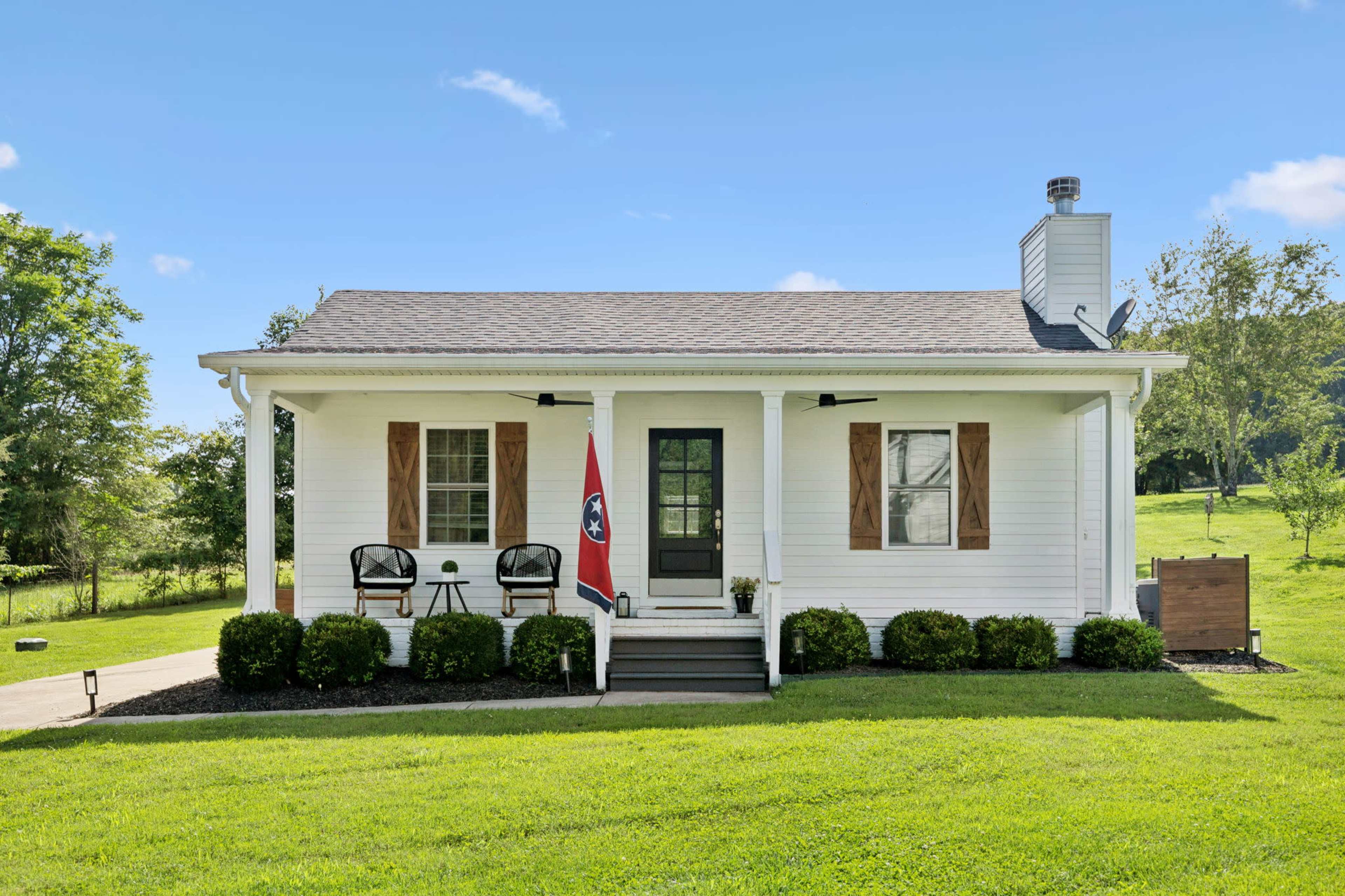 The image shows a single-story white house with a brown roof, featuring a small porch with two black chairs and a Tennessee flag.