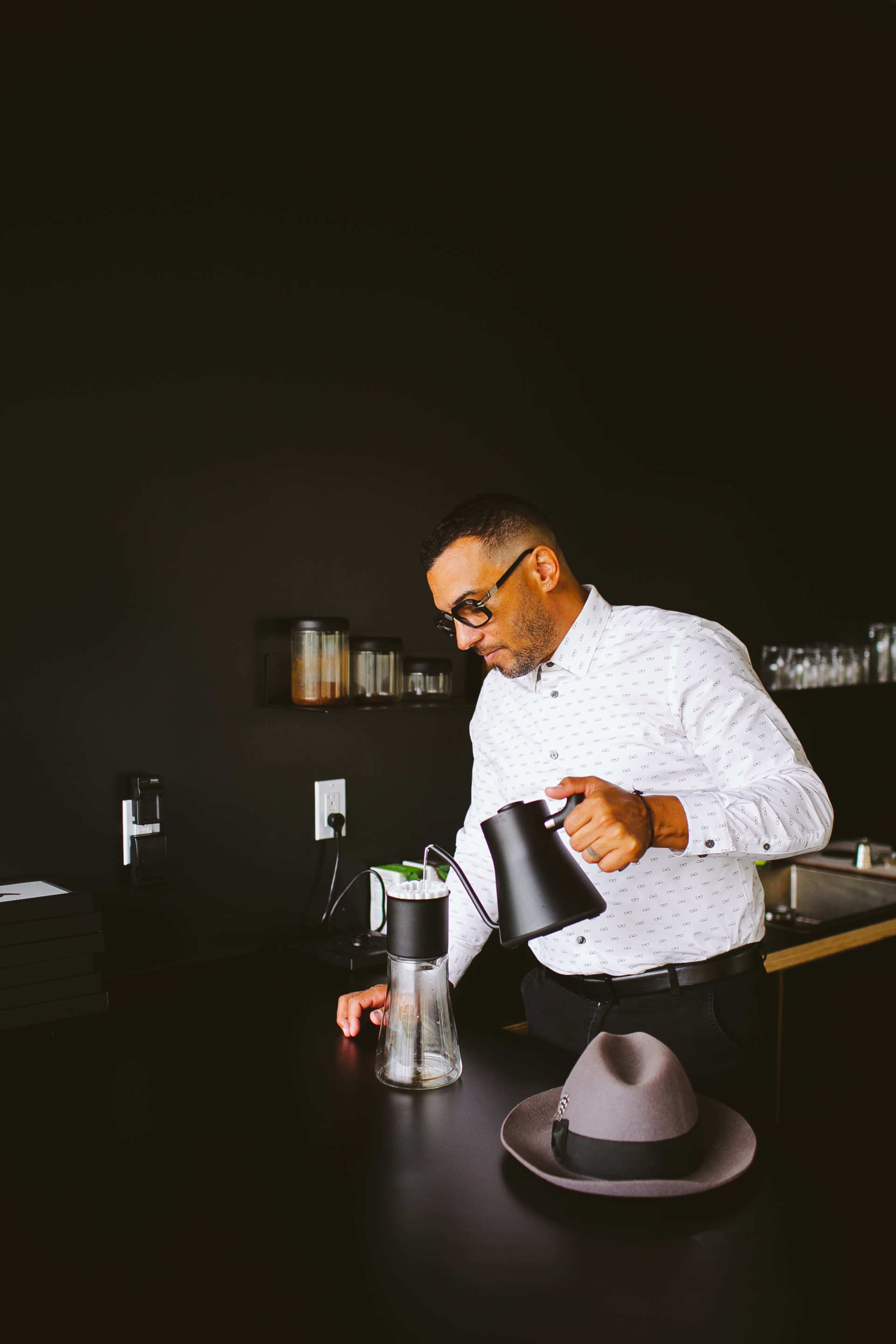 A man in a white shirt pours water from a black kettle into a glass container on a black table.