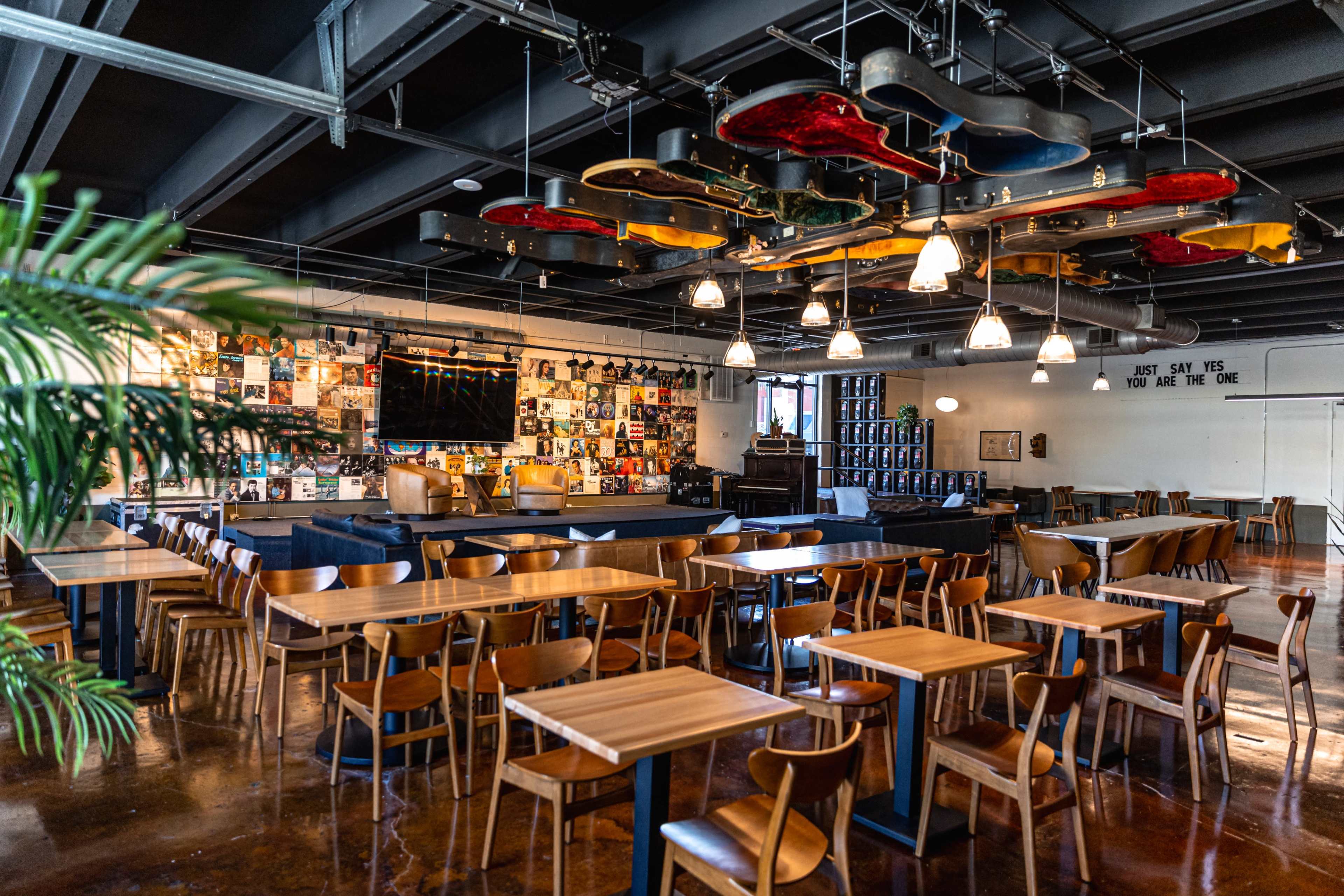 The image shows a modern dining area with wooden tables and chairs, featuring a wall decorated with records and a unique ceiling installation made of colorful guitars.
