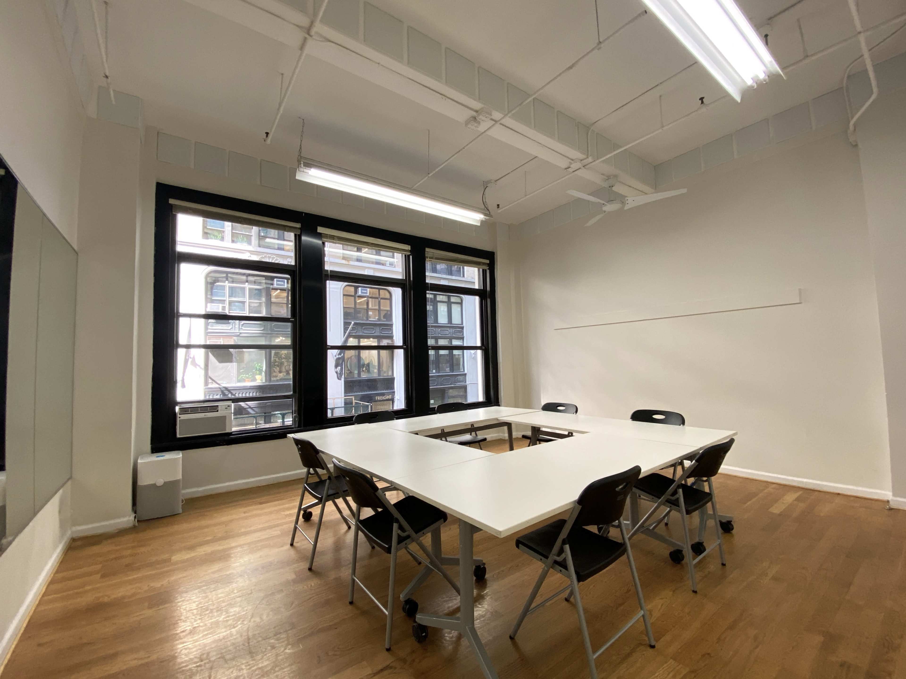 A spacious meeting room features a central table surrounded by black folding chairs and large windows that provide natural light.