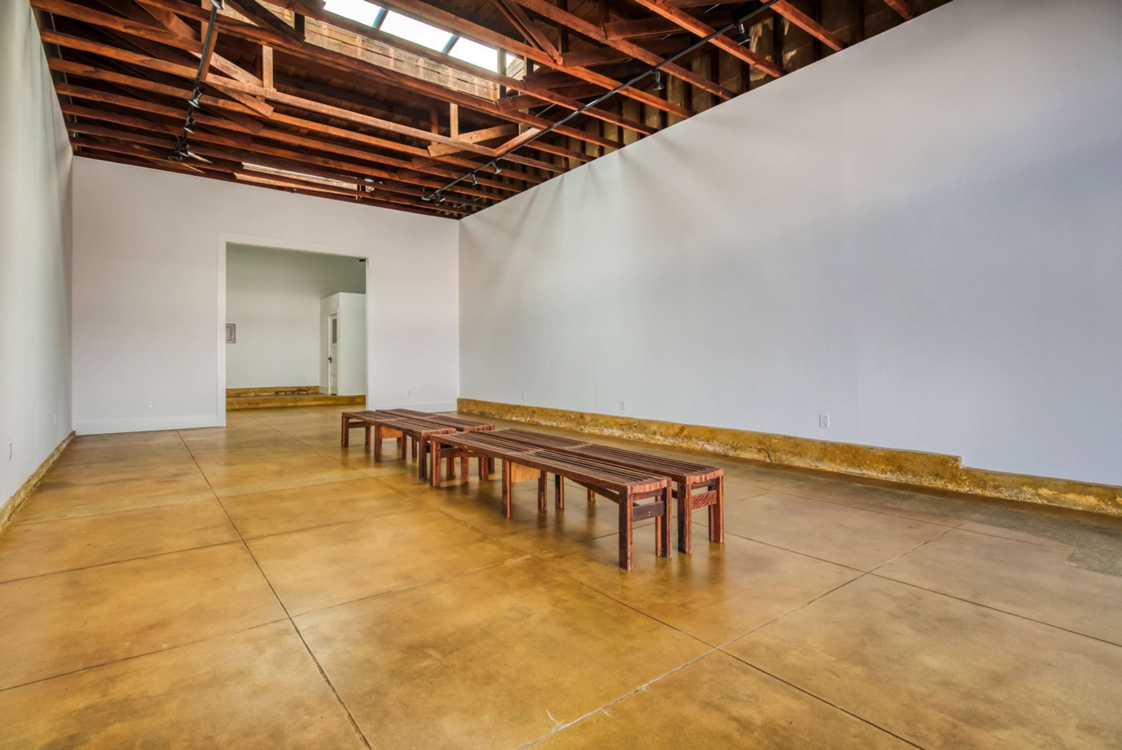 The image shows a spacious interior with exposed wooden beams, concrete floors, and several wooden benches arranged in a minimalist layout.