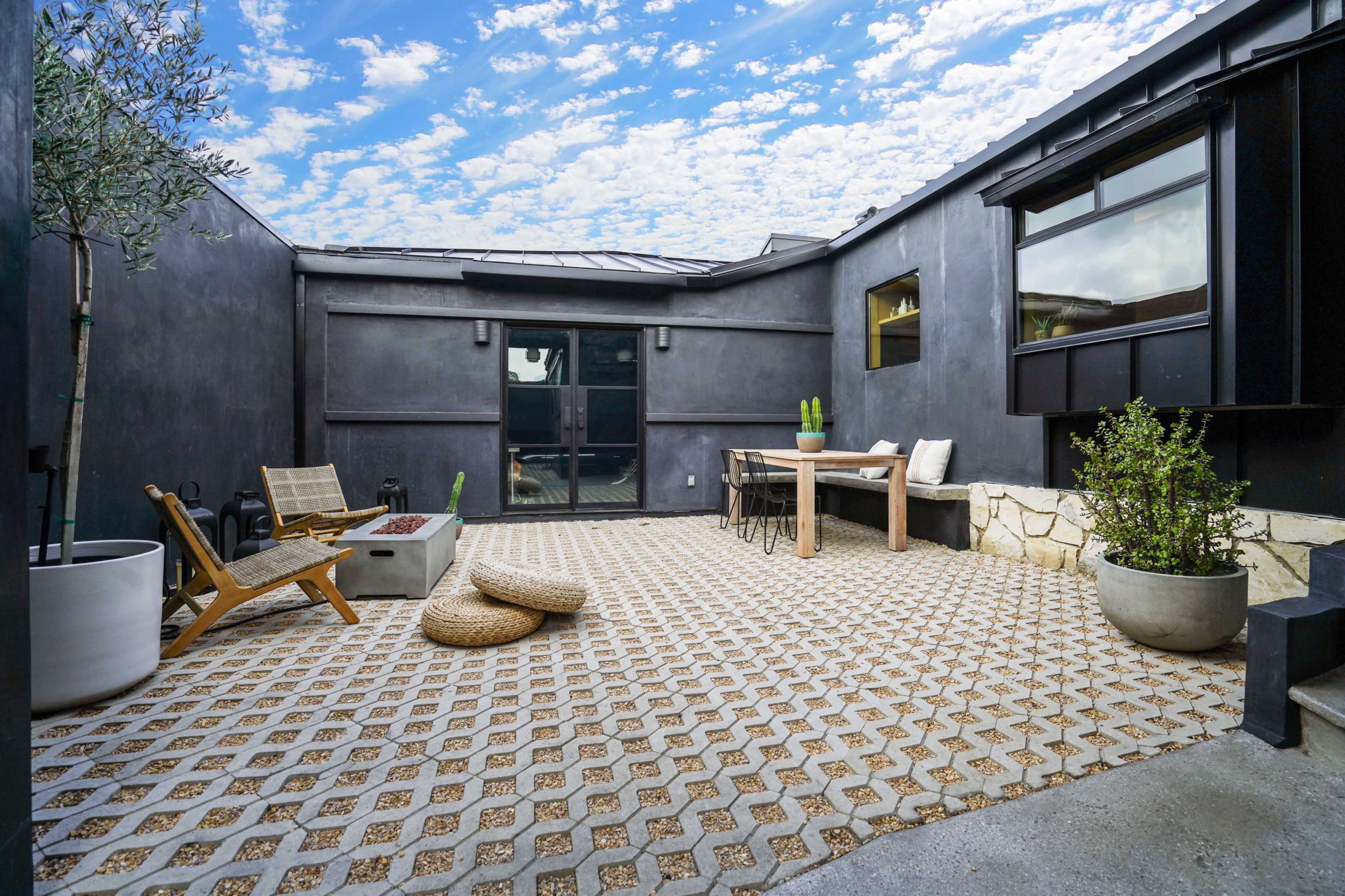 A modern, enclosed courtyard with a patterned stone floor, wooden furniture, and potted plants against black walls and a blue sky.