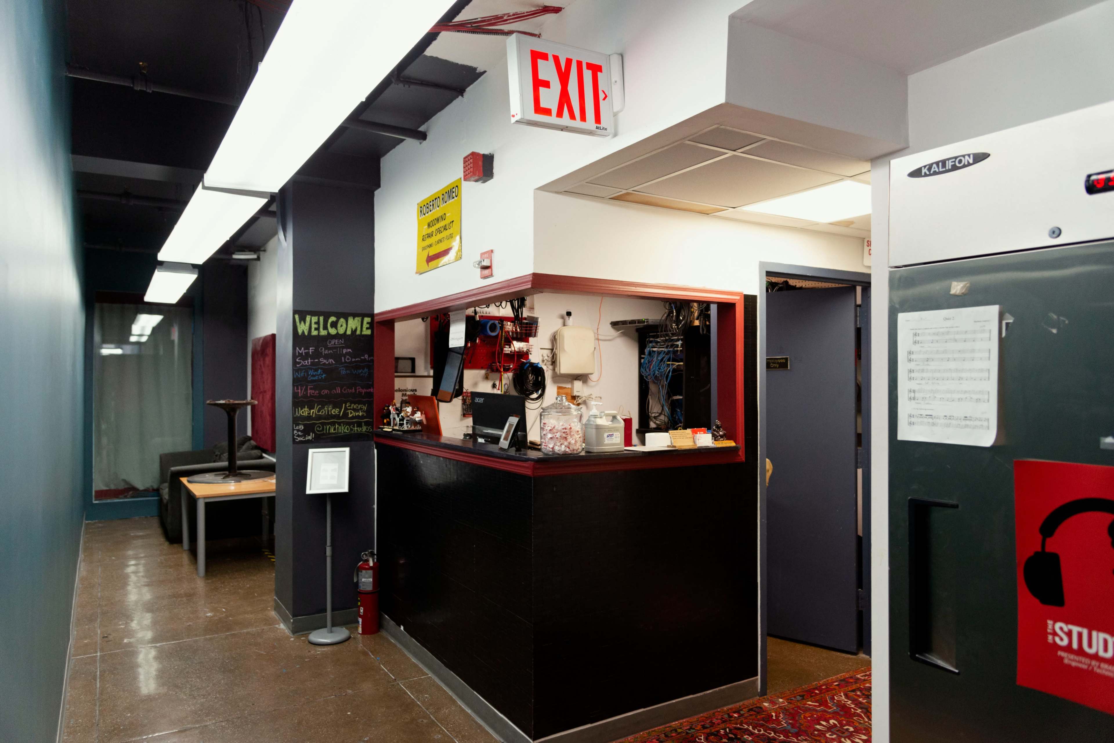 The image shows a corridor leading to a reception area with an "EXIT" sign, a chalkboard with welcoming text, and a counter with various items.