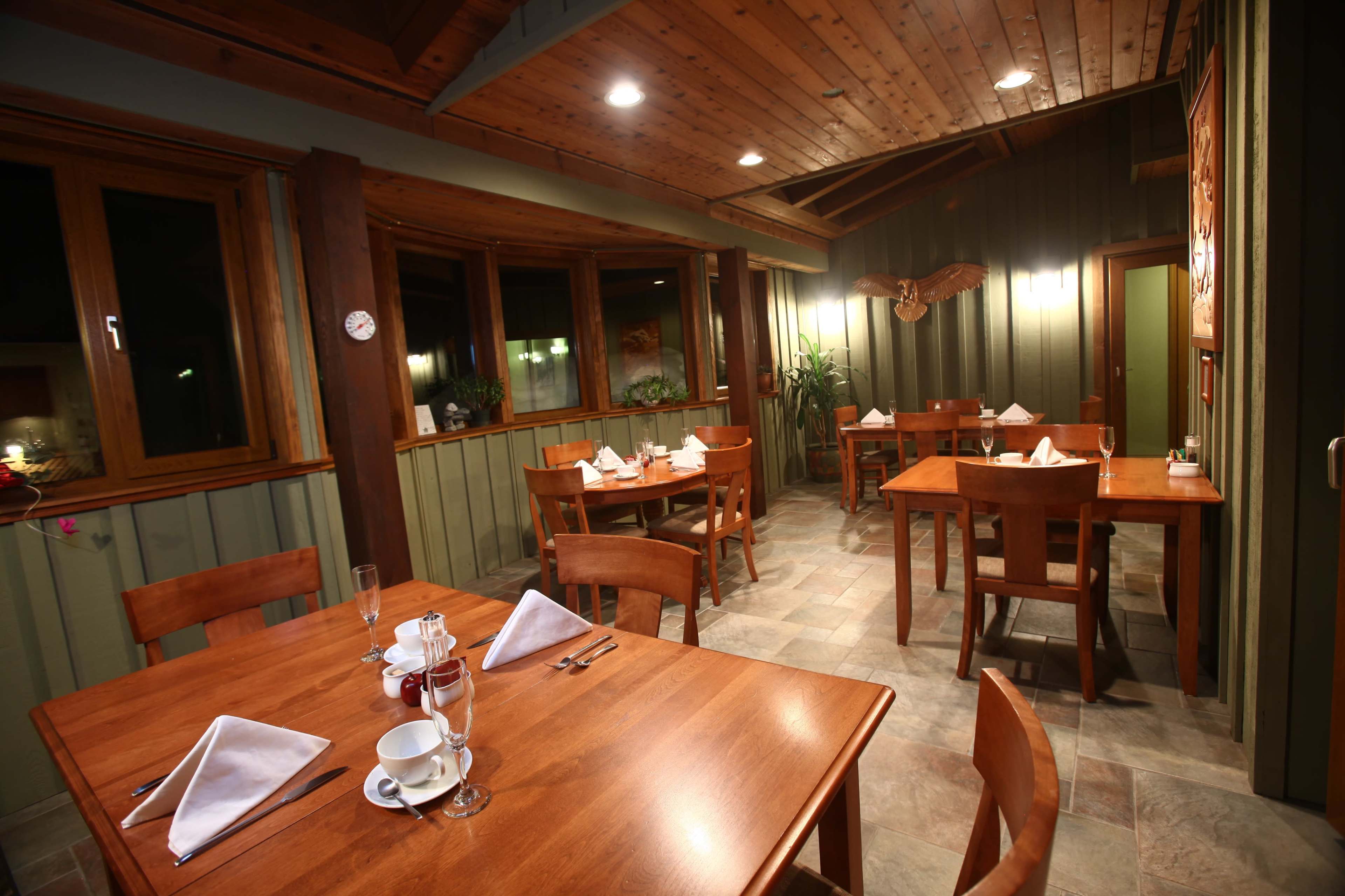 The image shows a cozy restaurant interior with wooden tables and chairs, set with white napkins and minimalist decor, illuminated by soft overhead lighting.