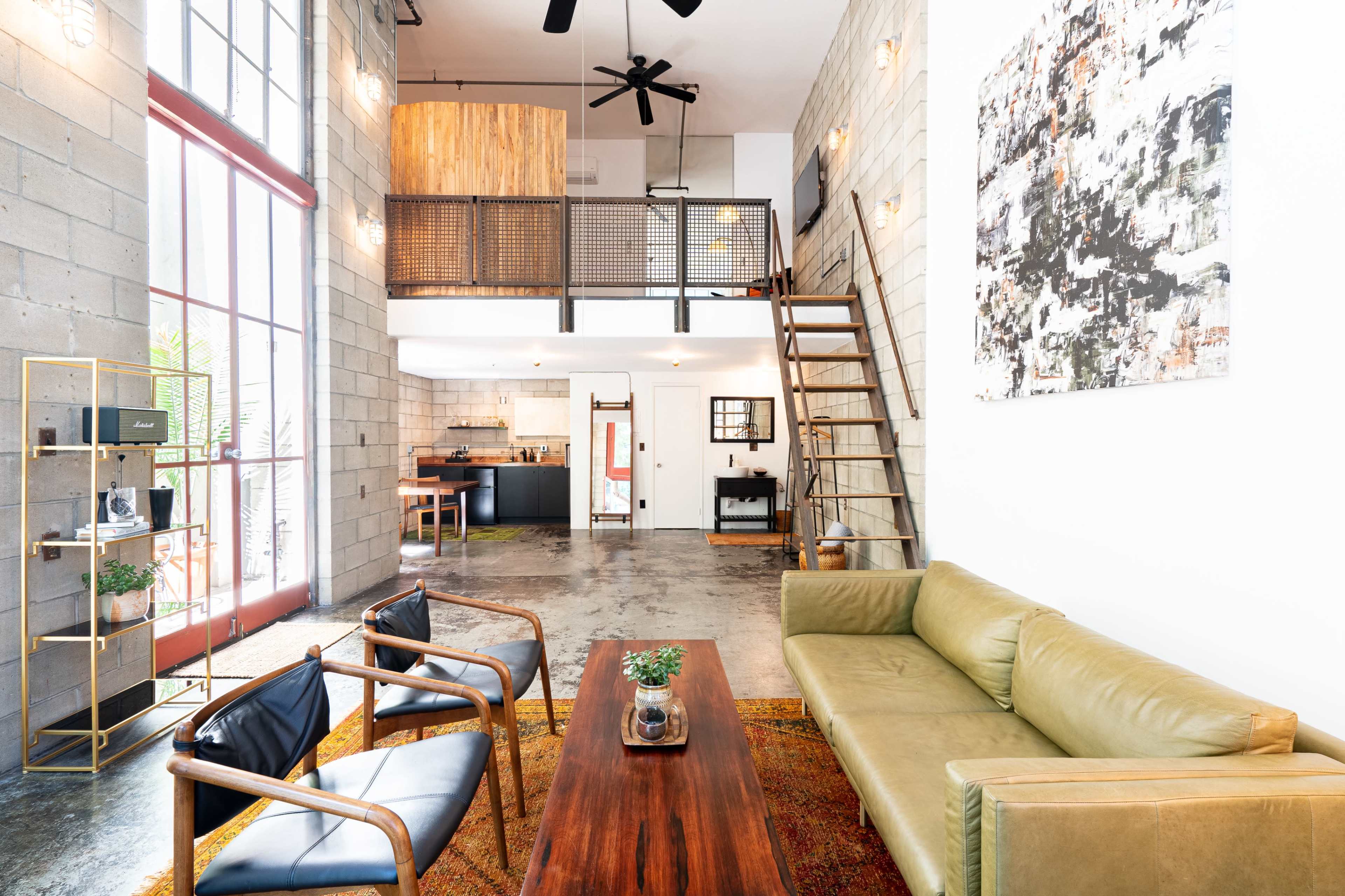 The image shows a modern loft-style living room with a green sofa, a wooden coffee table, and a staircase leading to a bedroom area.