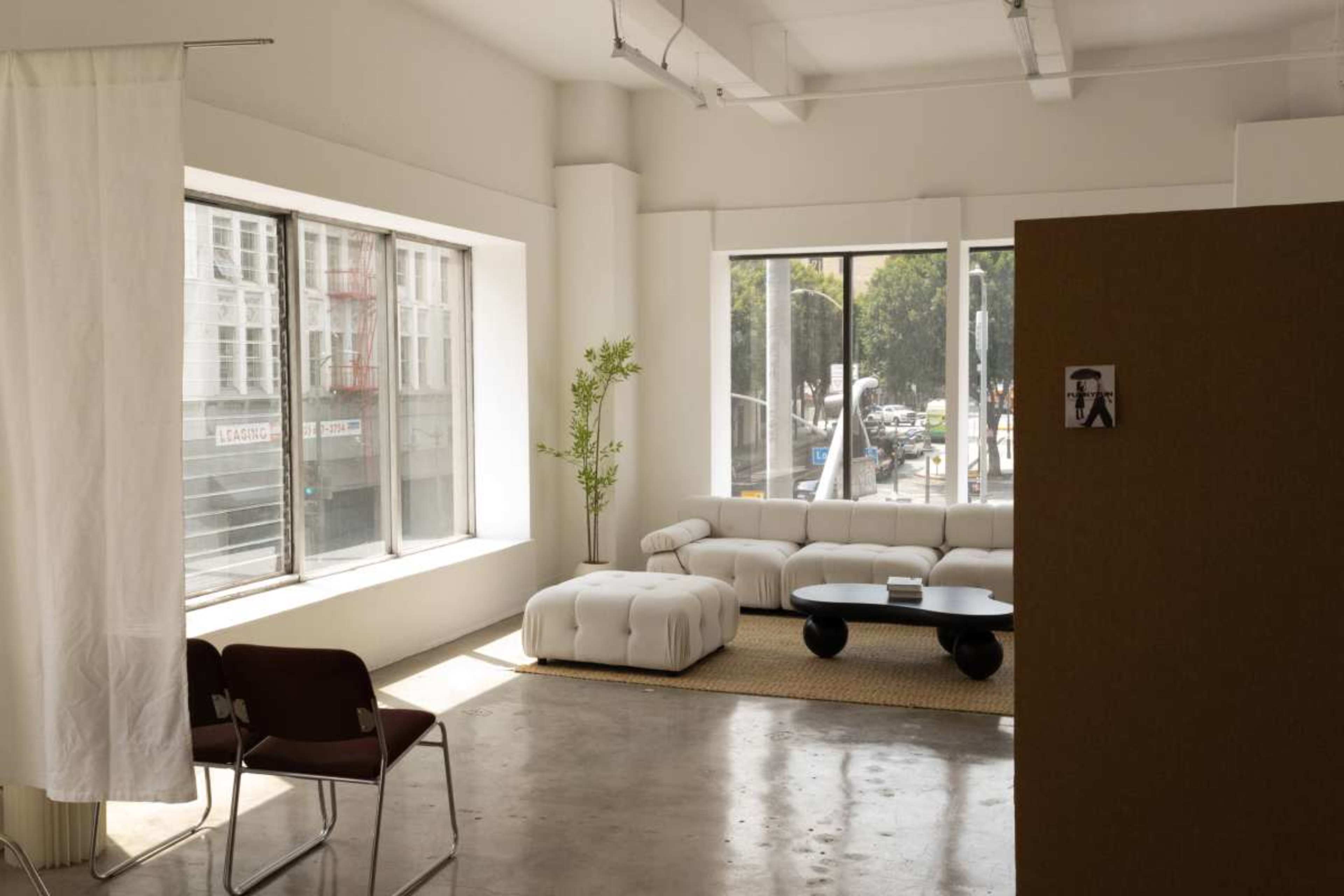 The image shows a minimalist interior with a white sectional sofa, a round black coffee table, and large windows letting in natural light.