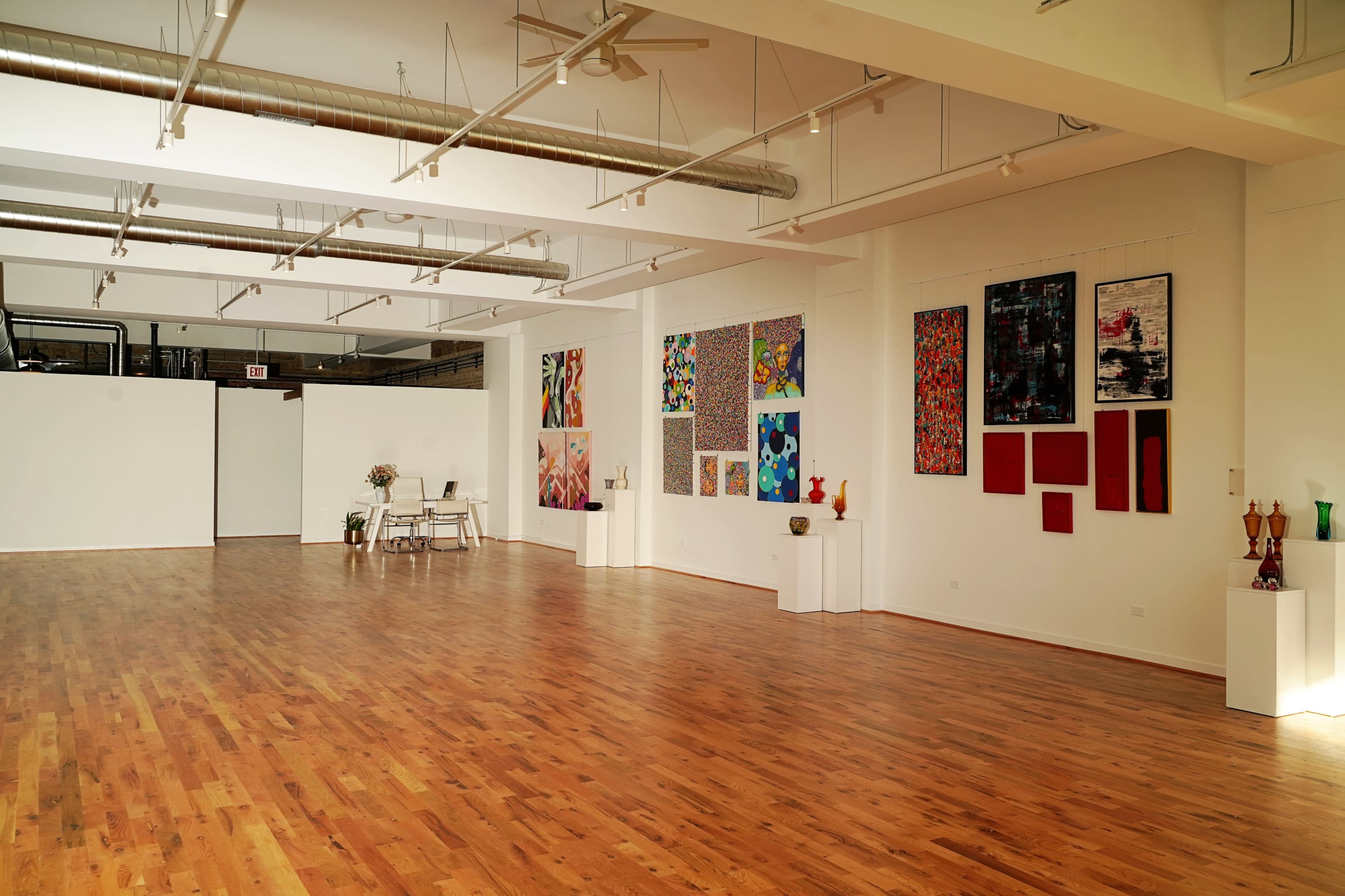 An art gallery features various colorful paintings on the walls and wooden flooring with a small seating area in the corner.