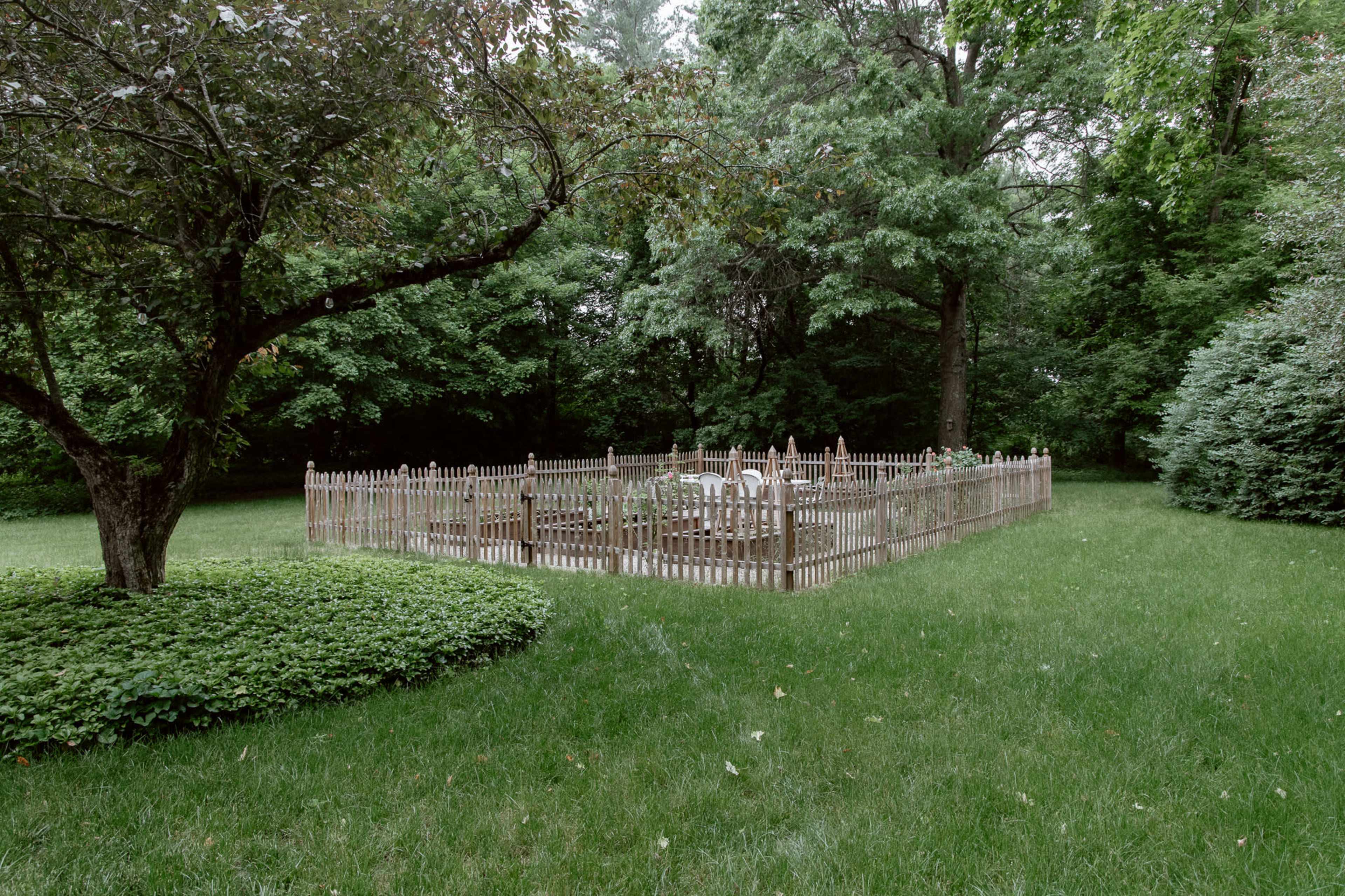 A wooden picket fence encloses a garden area surrounded by lush green grass and trees.