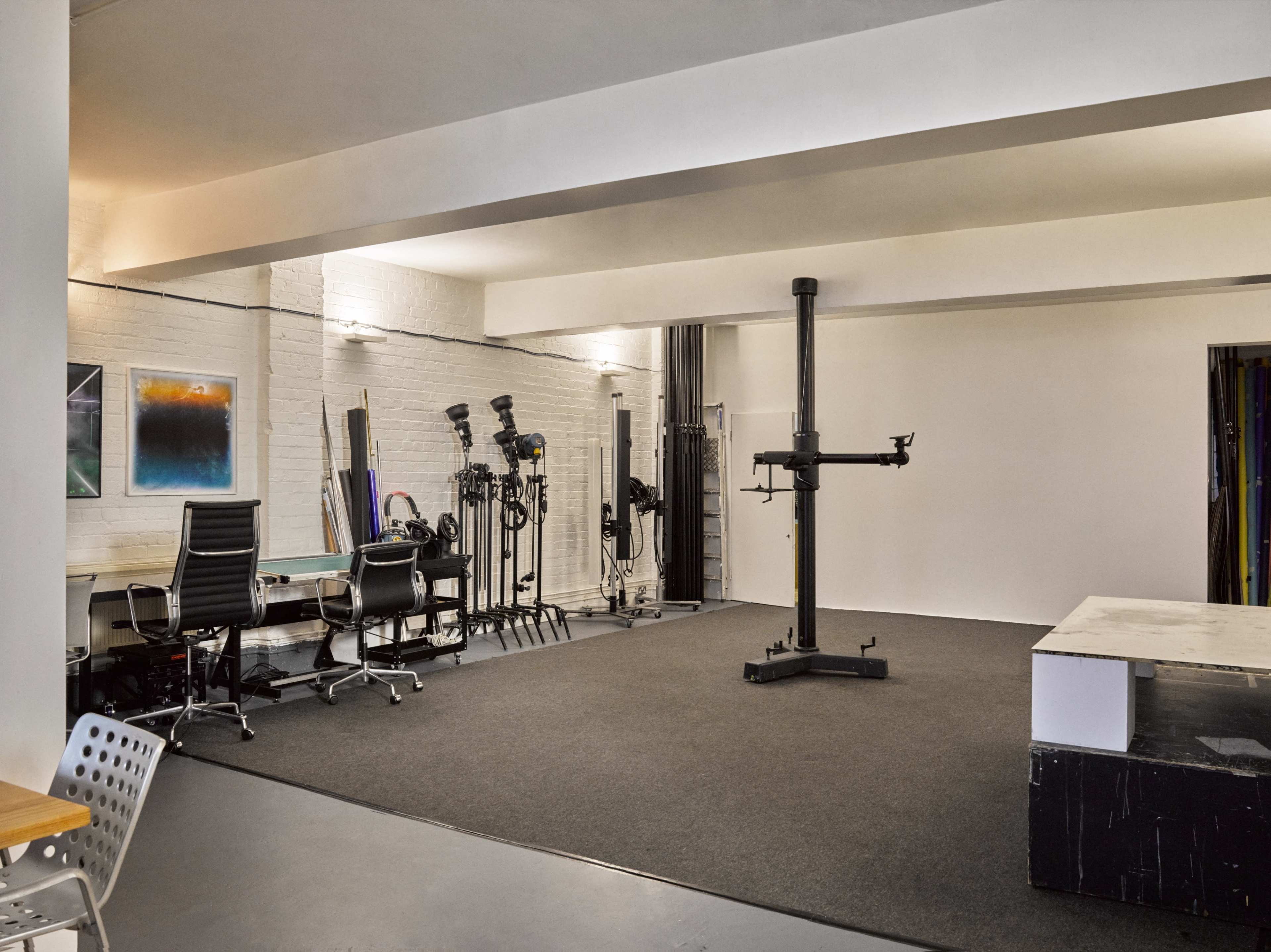 The image shows an empty studio space with a black carpet, two office chairs at a desk, several light stands, and a central equipment stand.