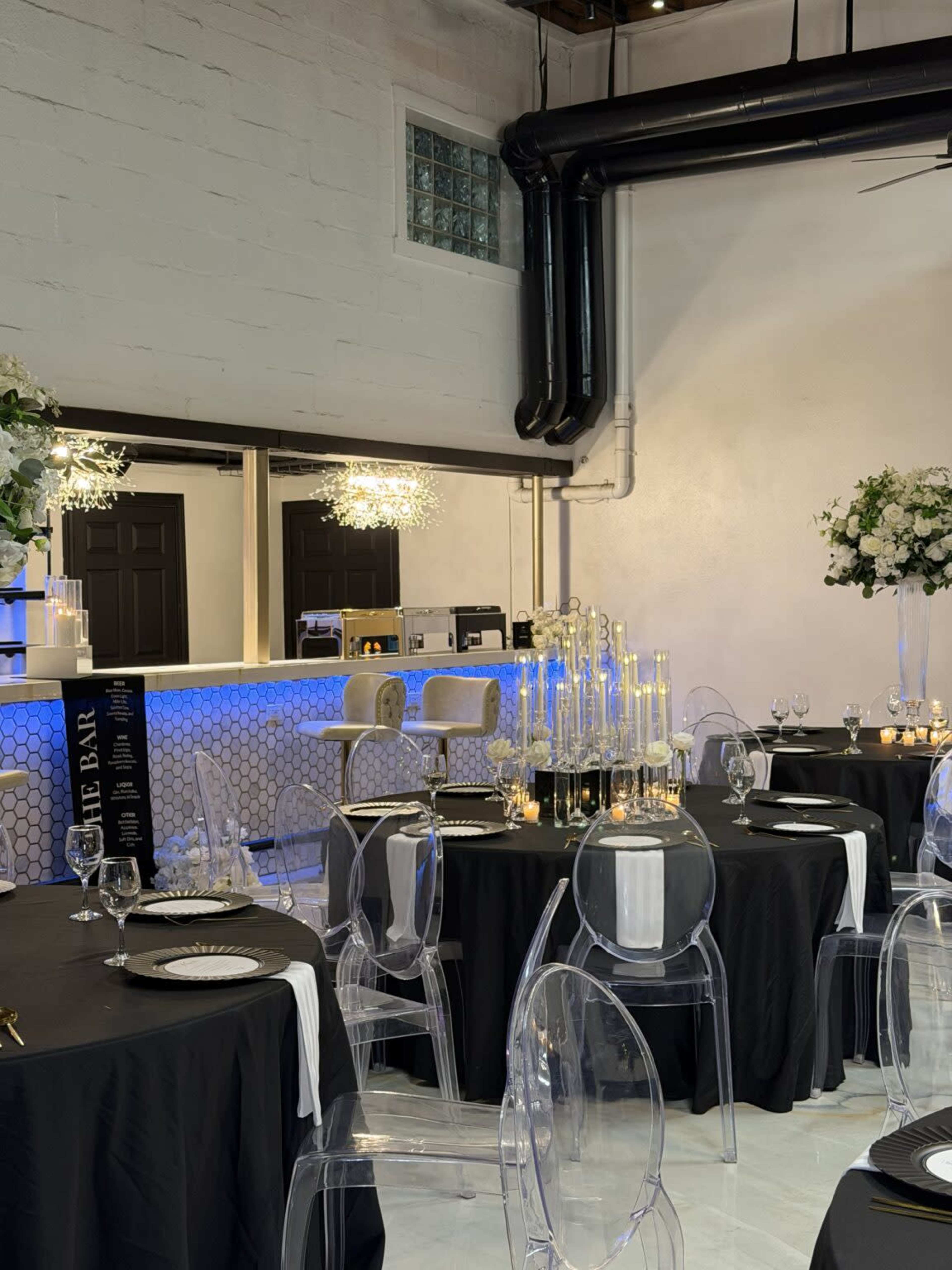 The image shows a modern event space with black tablecloths and clear acrylic chairs, featuring elegant centerpieces and a lit bar in the background.