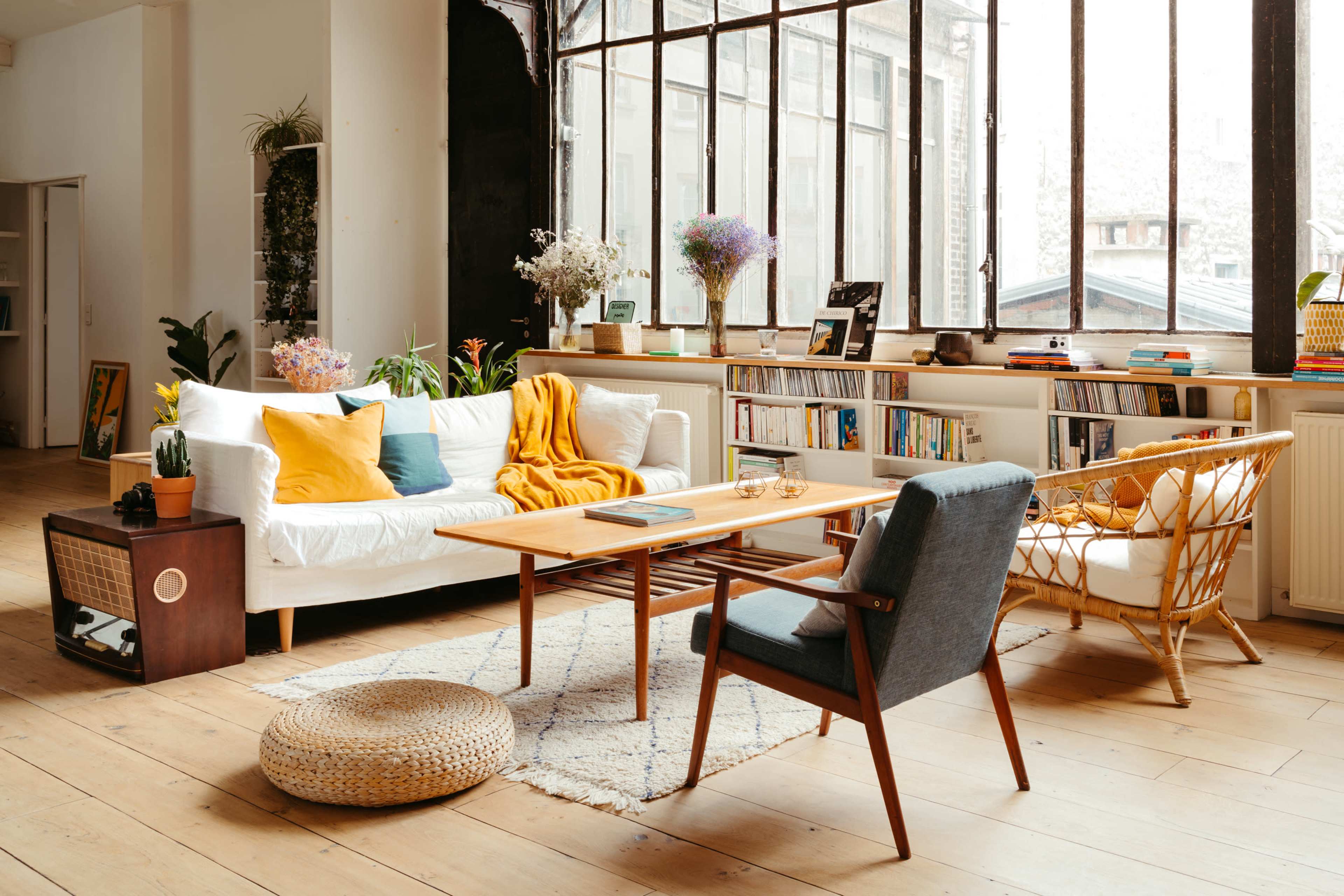 A bright living room features a white sofa with colorful cushions, a wooden coffee table, and large windows that allow natural light to fill the space.