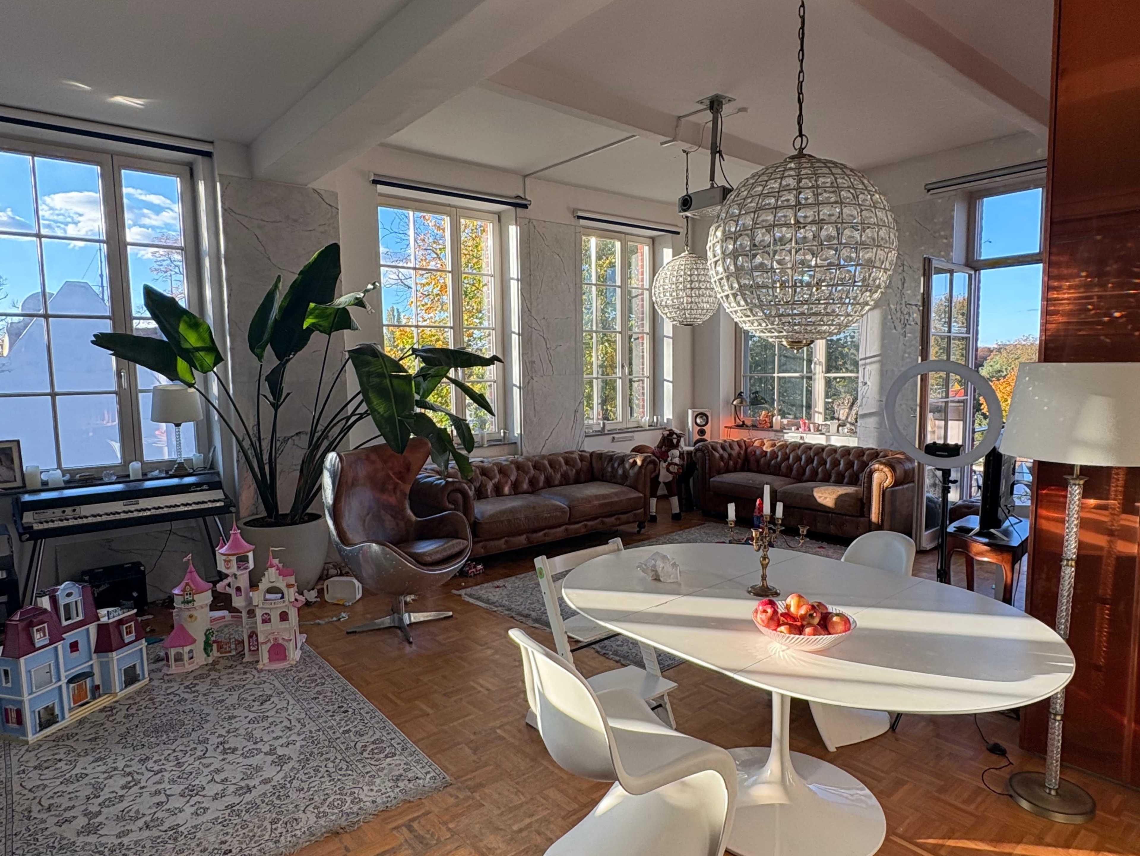 A bright living space features a white dining table surrounded by modern chairs, leather sofas, large windows, and decorative plants.