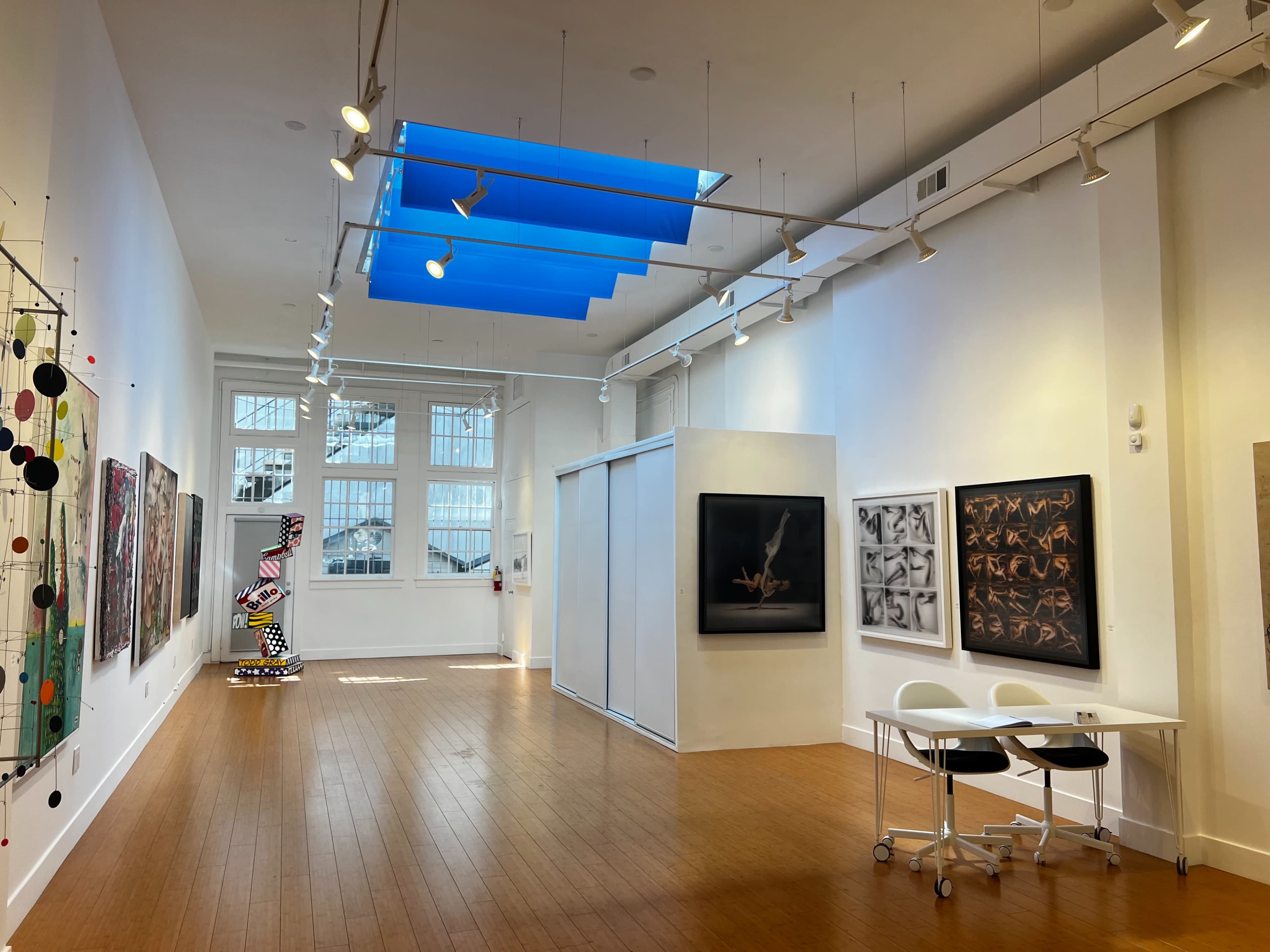 The image shows a spacious art gallery with various artworks displayed on the walls and a sculptural piece in the center, illuminated by natural light through a skylight.