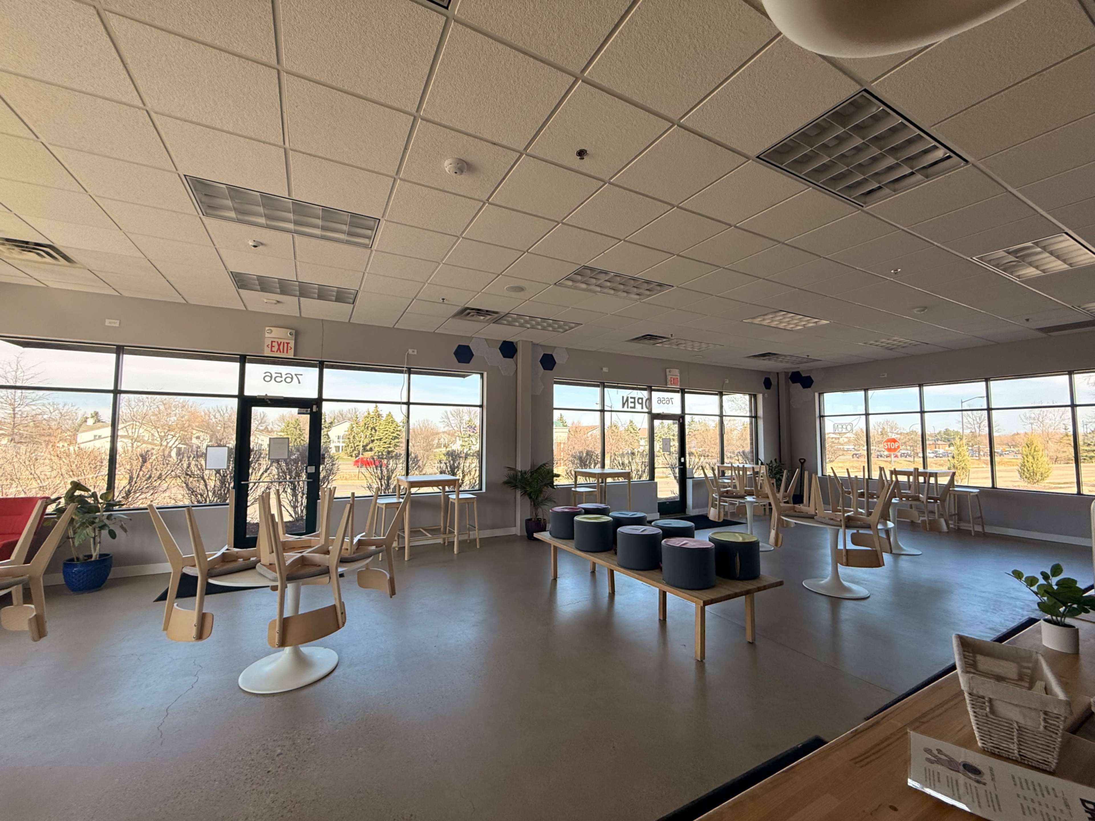The image shows a spacious, modern cafe with tables and chairs stacked upside down, large windows letting in natural light, and a few plants arranged around the room.