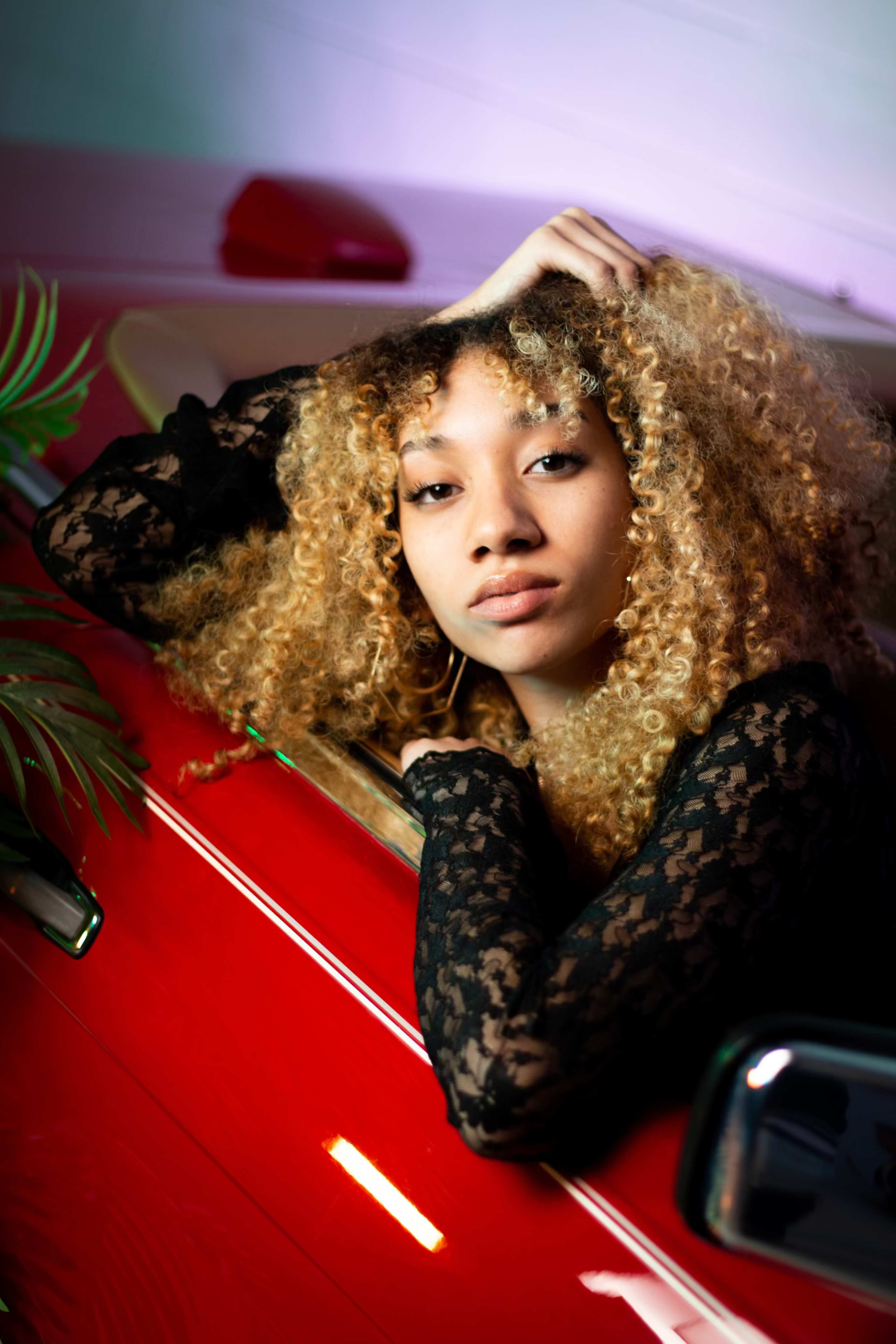 A person with curly hair rests their arm on the edge of a red car while looking directly at the camera.