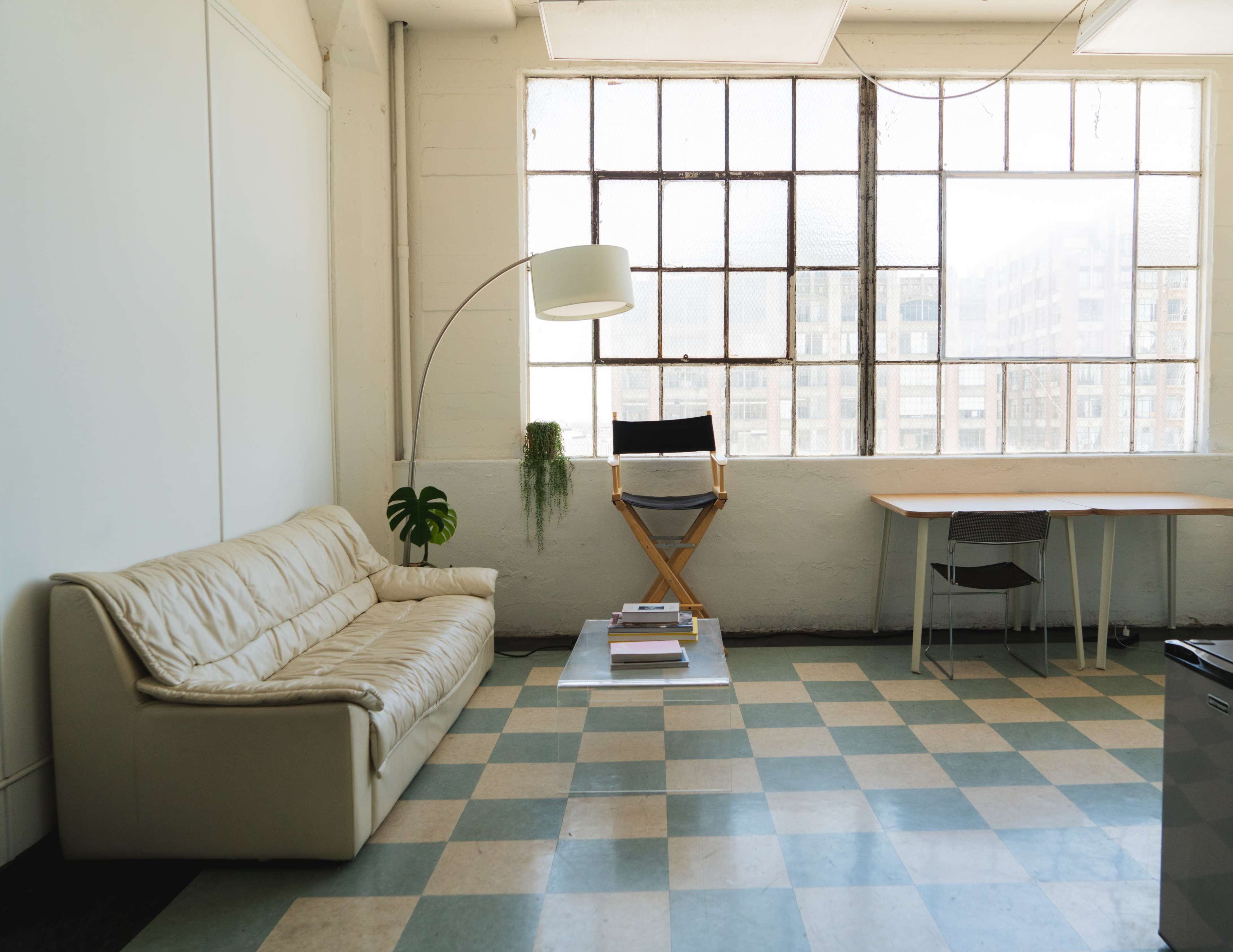 The image shows a spacious room with a leather couch, a tall lamp, a folding chair by a large window, and a simple wooden table on a checkered floor.