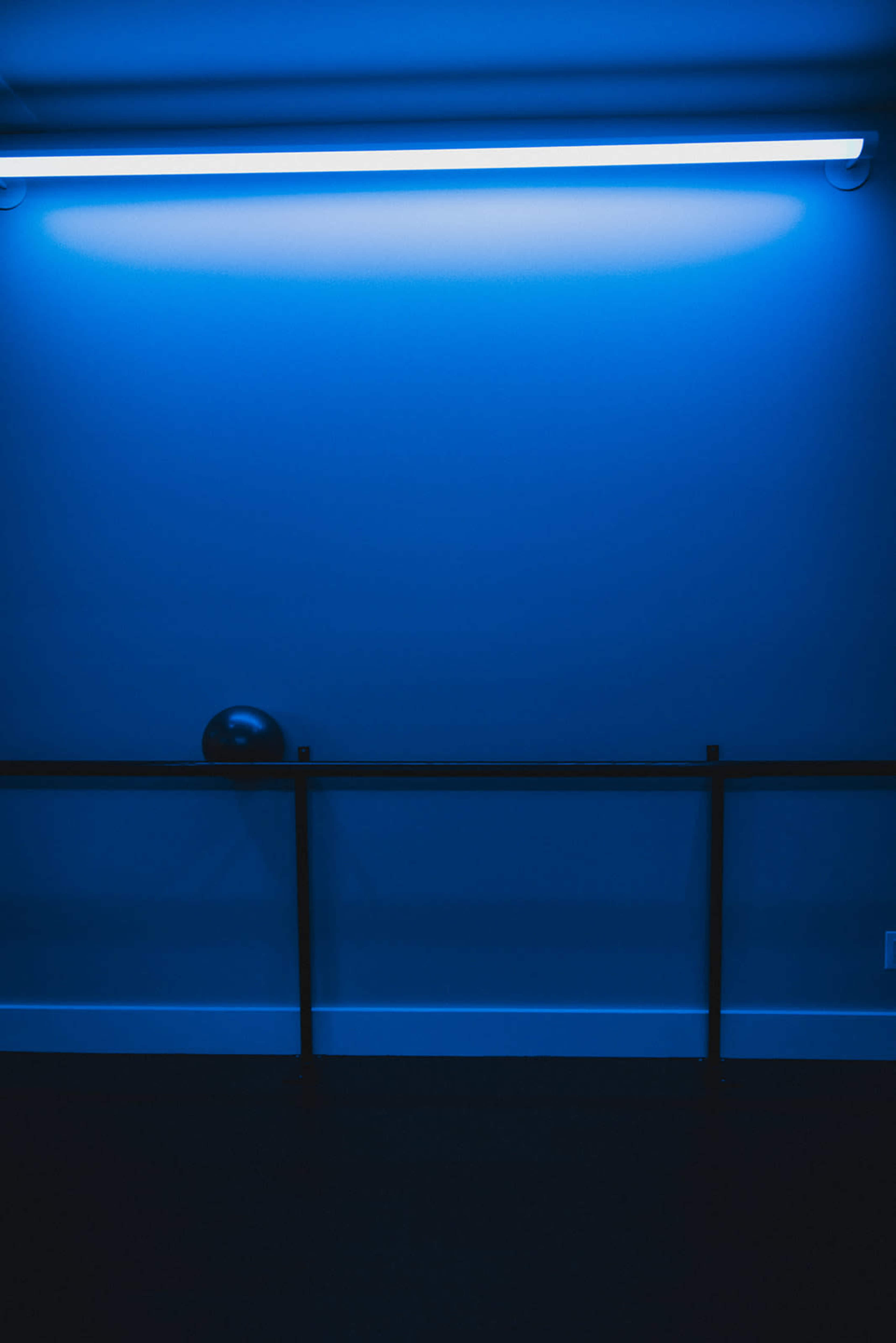 A black sphere sits on a railing beneath a blue-lit wall.
