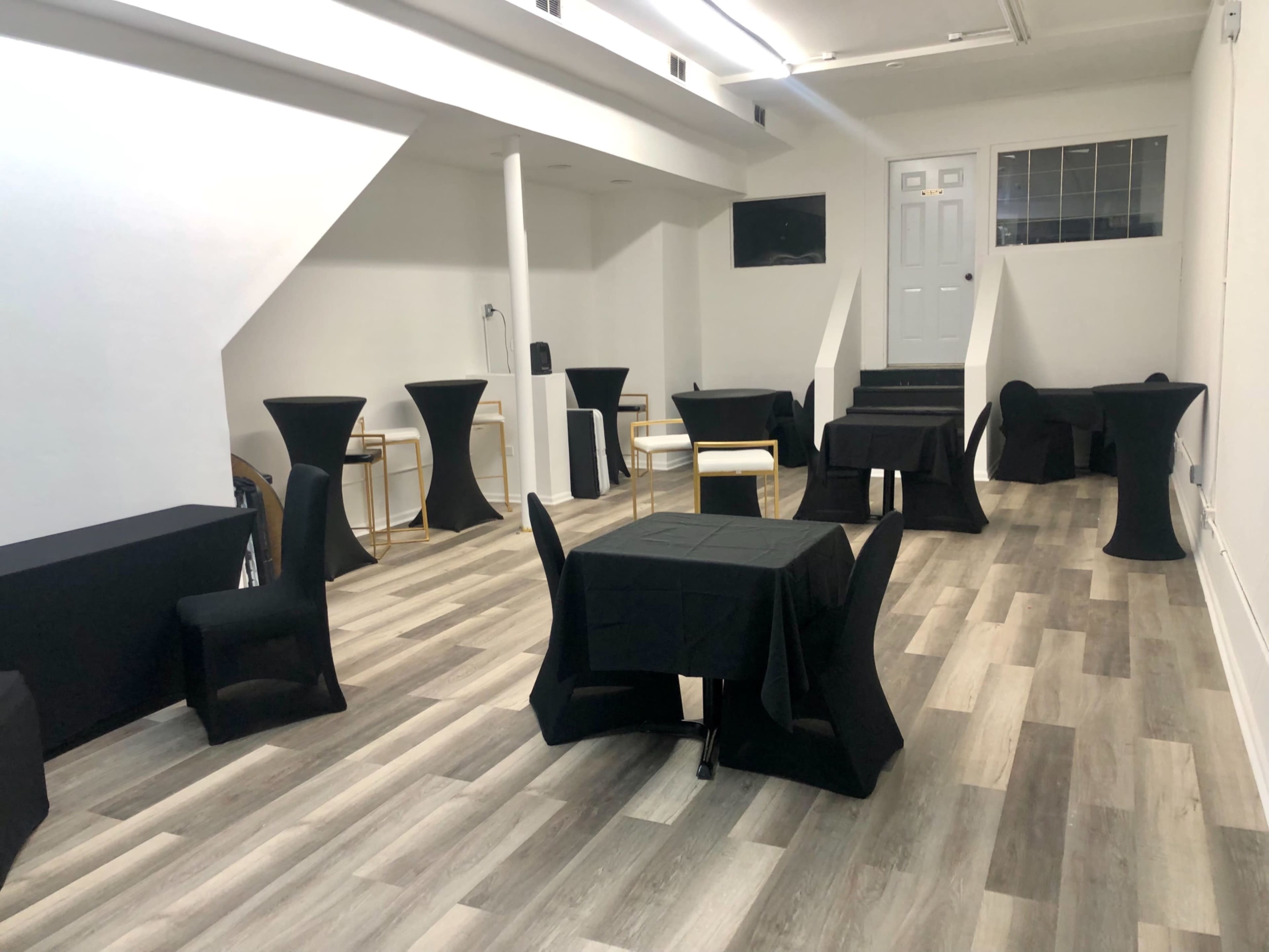 The image shows a minimalist event space with several black tables and chairs, arranged on a light wooden floor, leading to a stairway and a door at the rear.