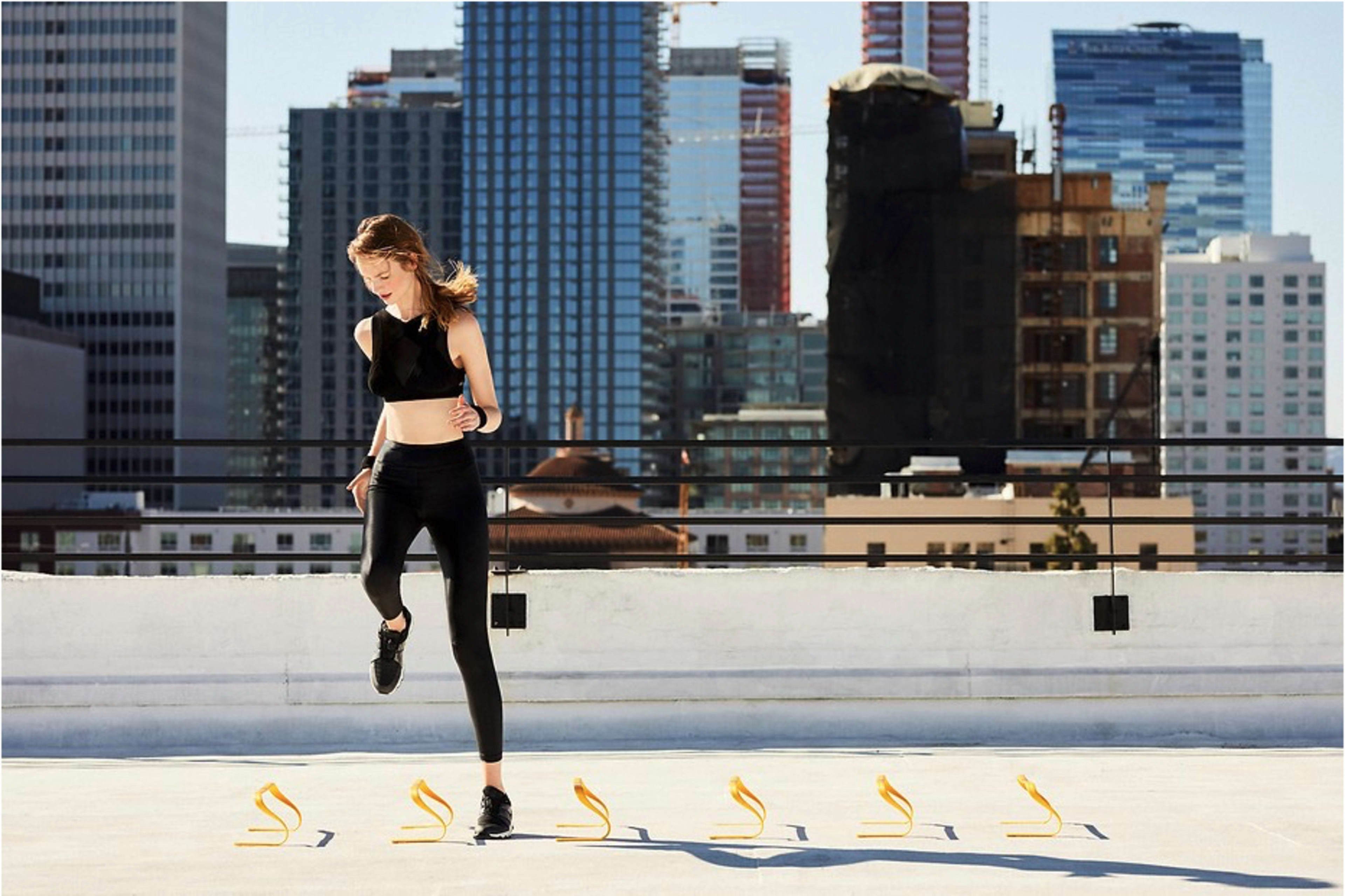 A woman in athletic wear performs agility drills using hurdles on a rooftop with a city skyline in the background.