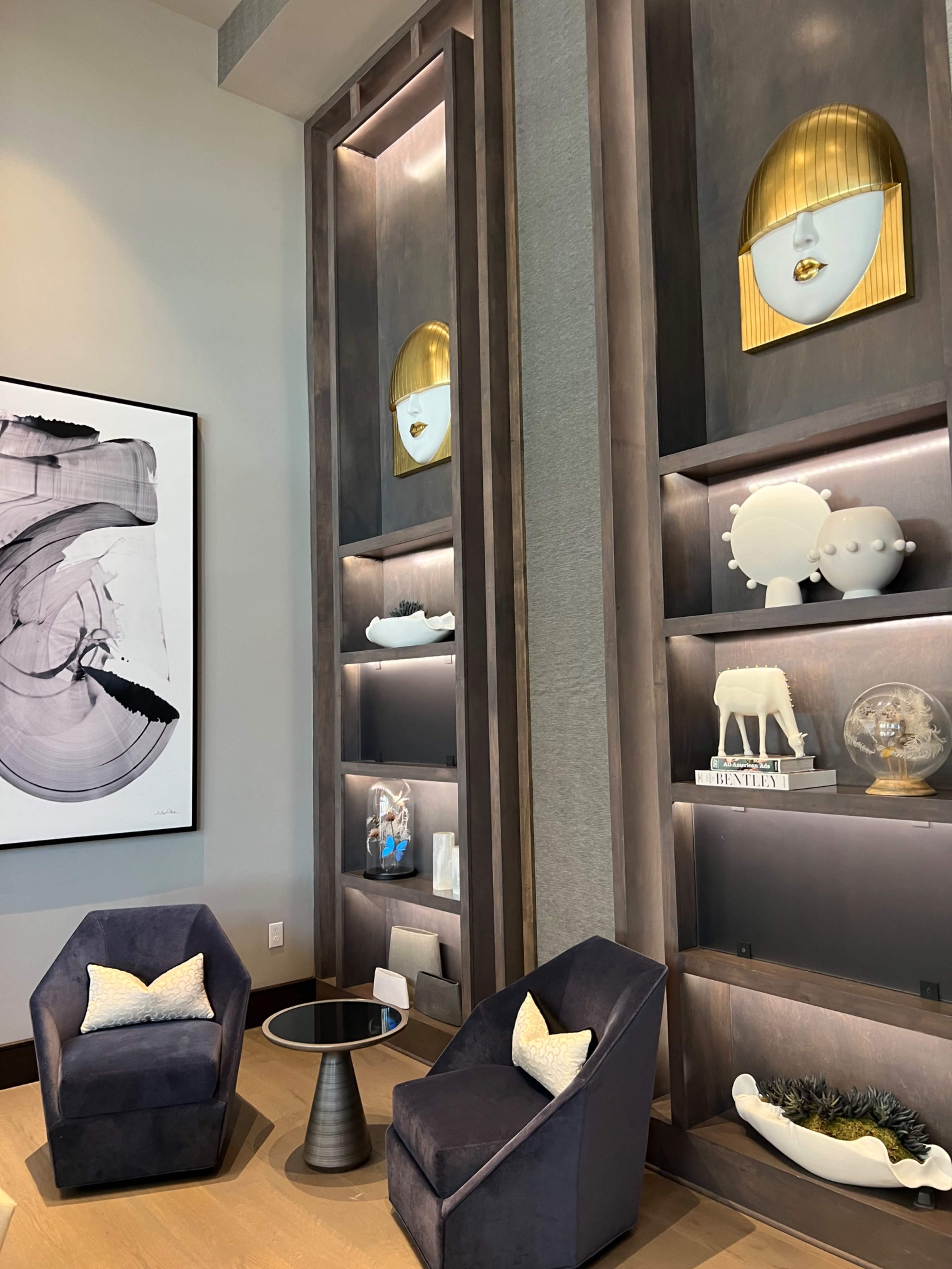 A modern interior with two velvet armchairs, a round table, and a decorative shelving unit displaying art and collectibles.