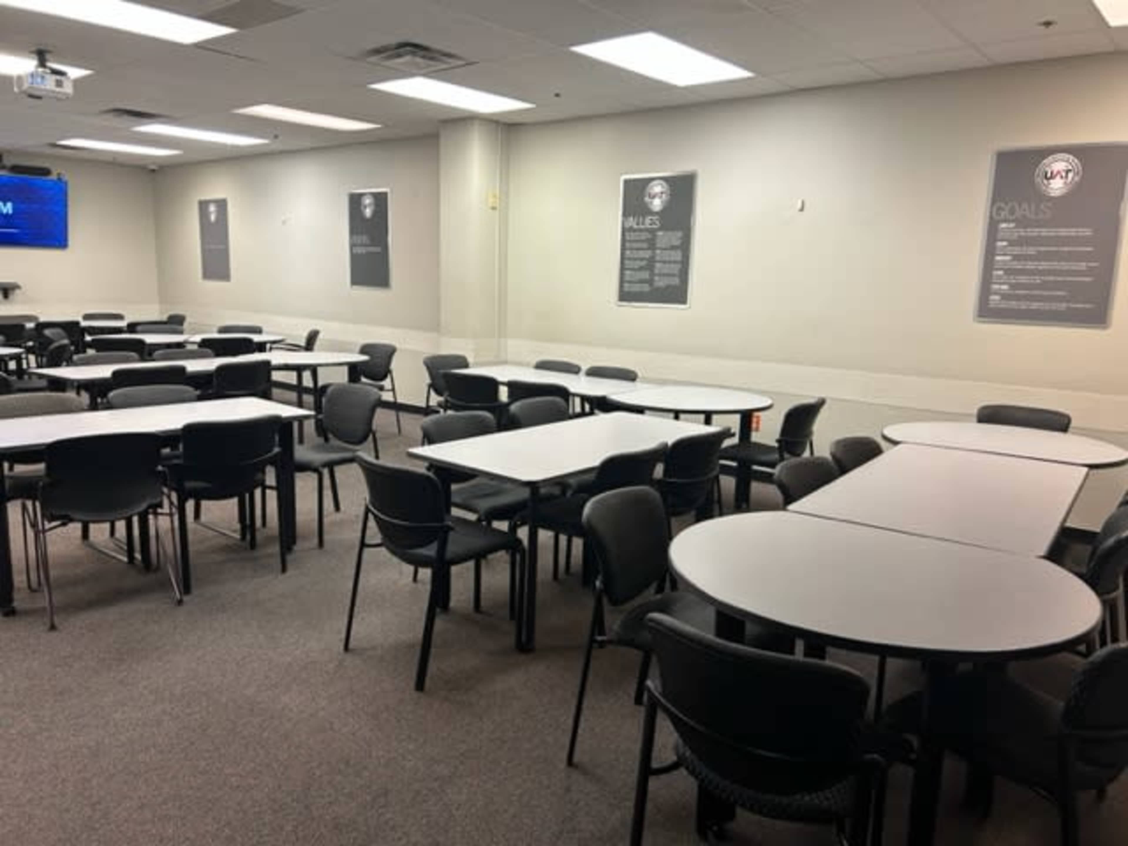 A group of round and rectangular tables surrounded by black chairs is arranged in a room with beige walls and motivational posters.