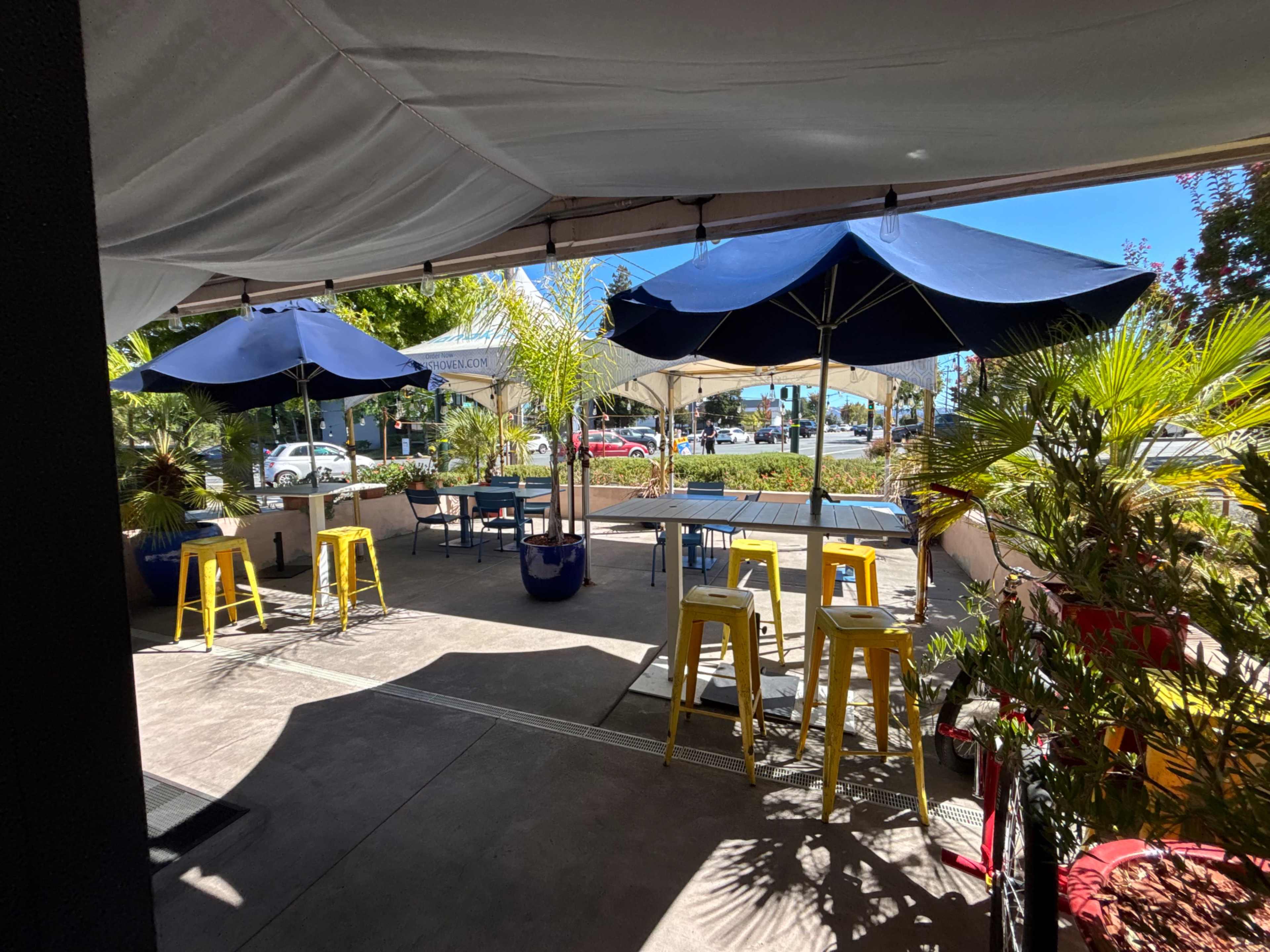 The image shows an outdoor patio area with blue umbrellas, yellow stools, and potted plants under a white canopy.
