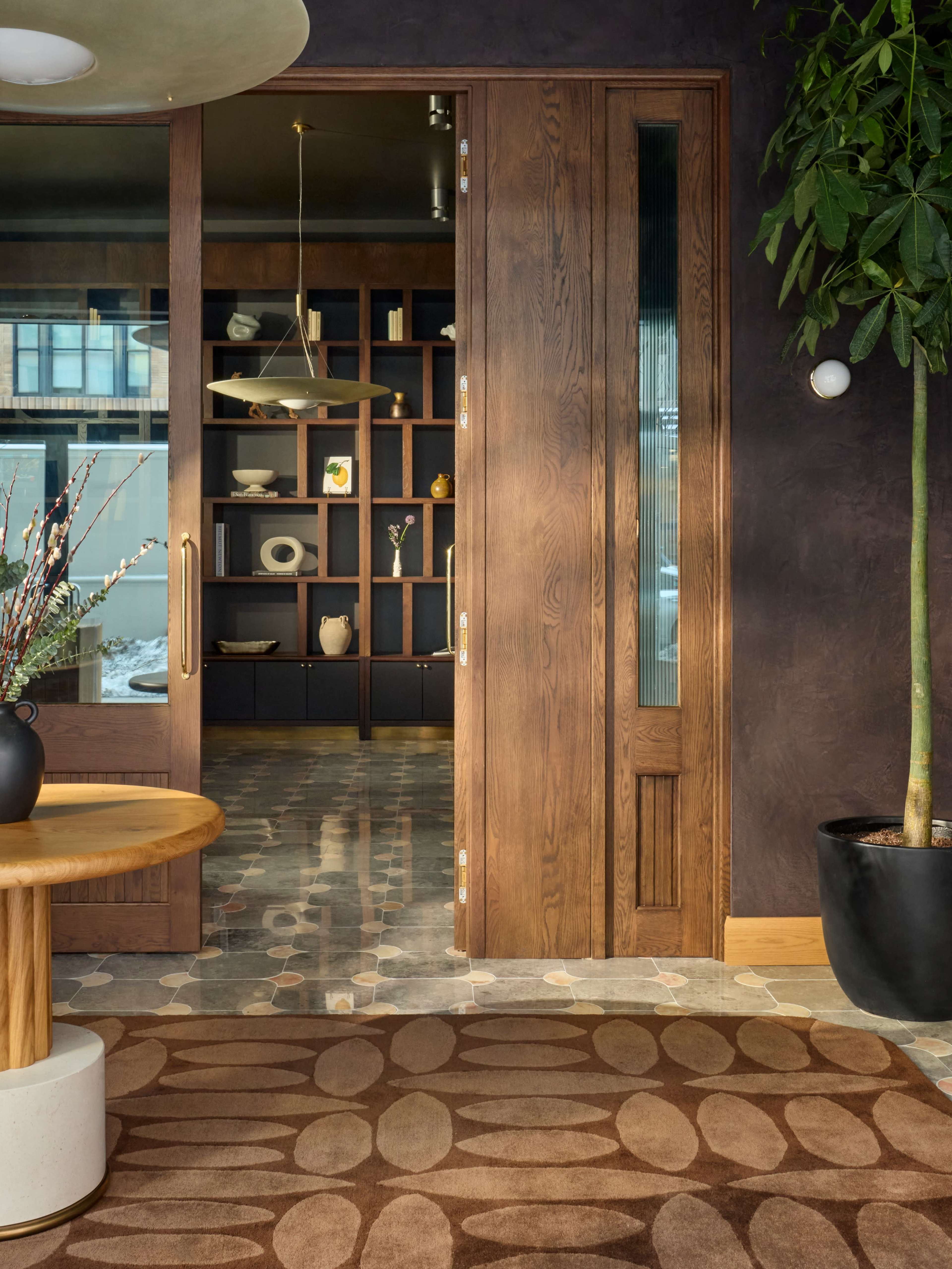 A modern entrance features a wooden door that opens to a stylish interior with geometric shelves and decorative items, complemented by greenery and a round table.