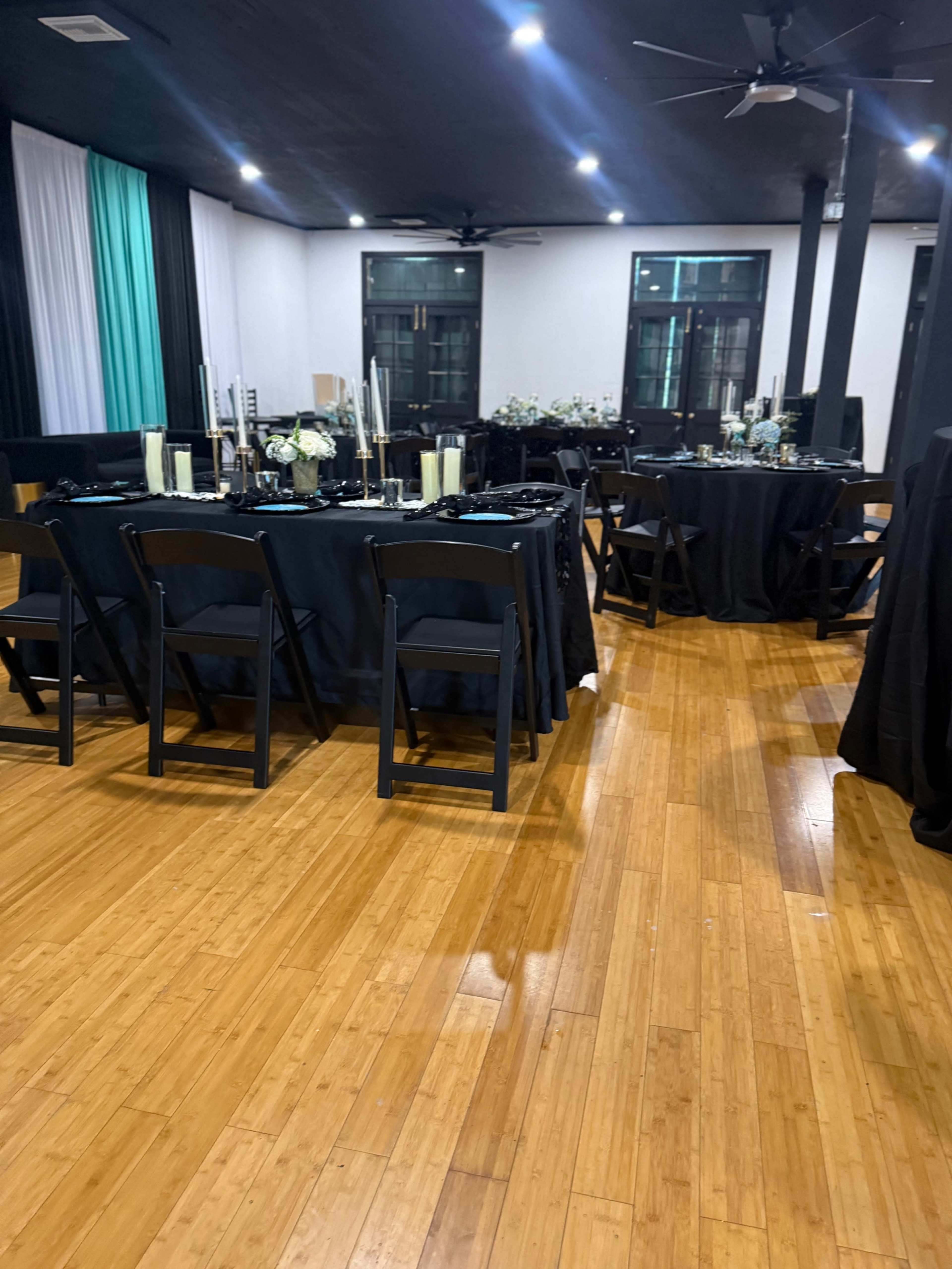 The image shows a spacious event venue with black dining tables set with elegant centerpieces, surrounded by wooden flooring and soft lighting.