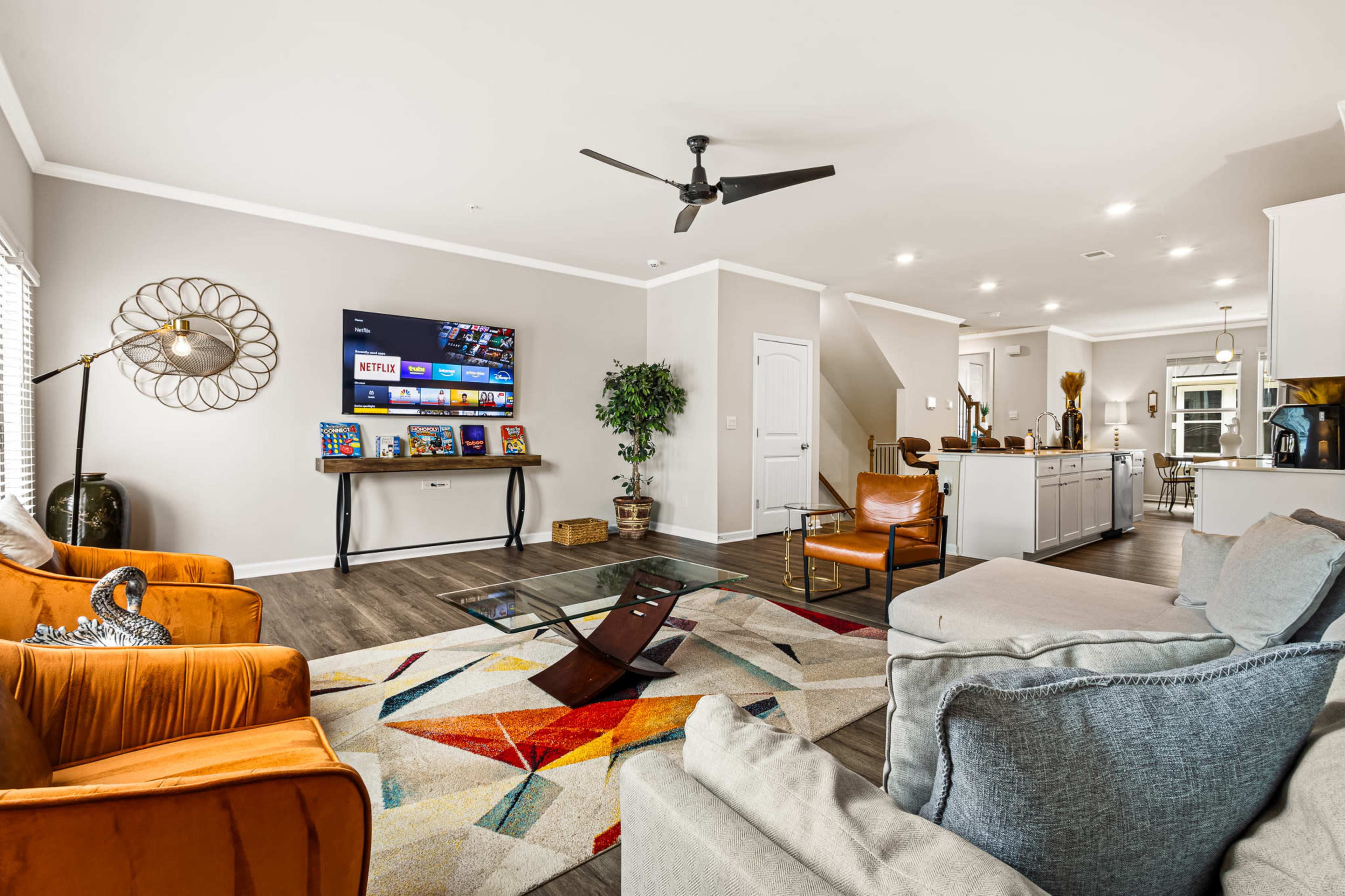 The living room features an array of seating options, a wall-mounted television displaying streaming services, and a kitchen visible in the background.