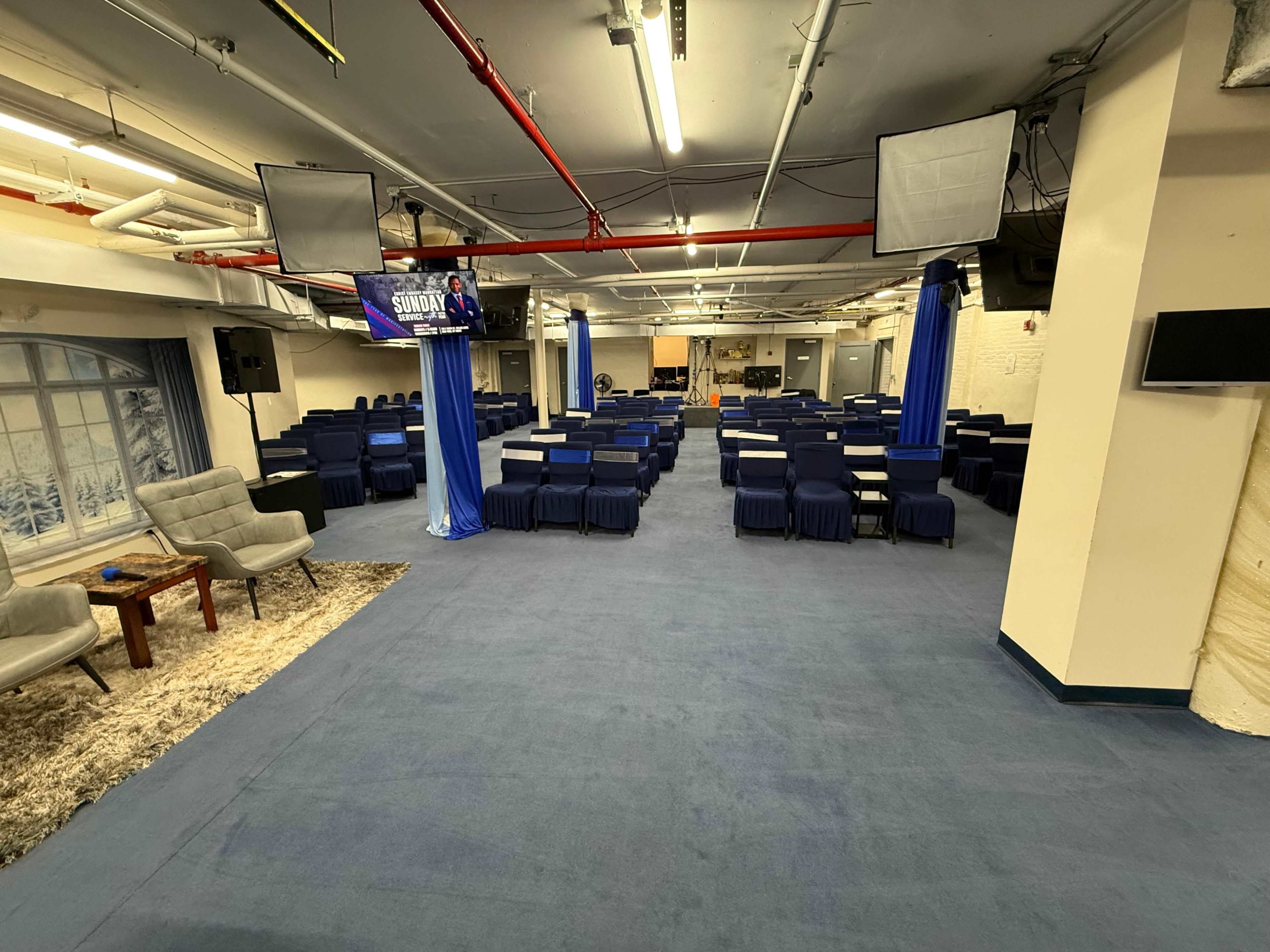 The image shows a spacious indoor venue arranged with rows of blue chairs and a few tables, with screens mounted on the walls.