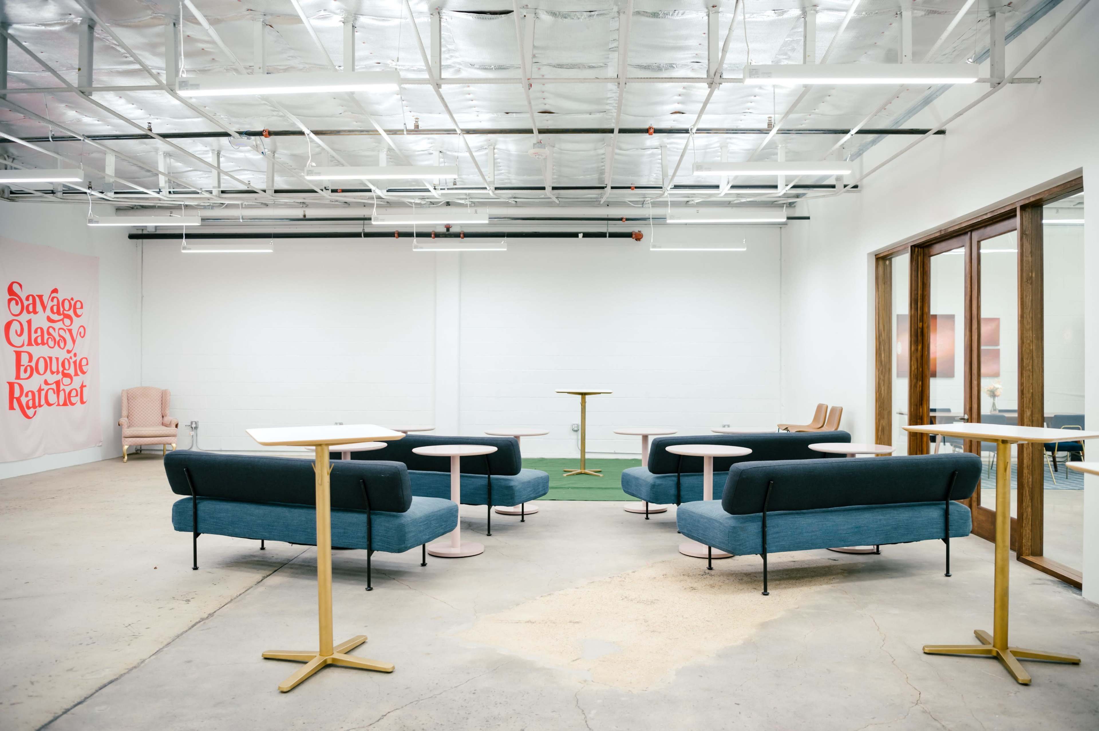 The image shows a modern, minimalist lounge area with several teal sofas and small round tables in a spacious, brightly lit room.