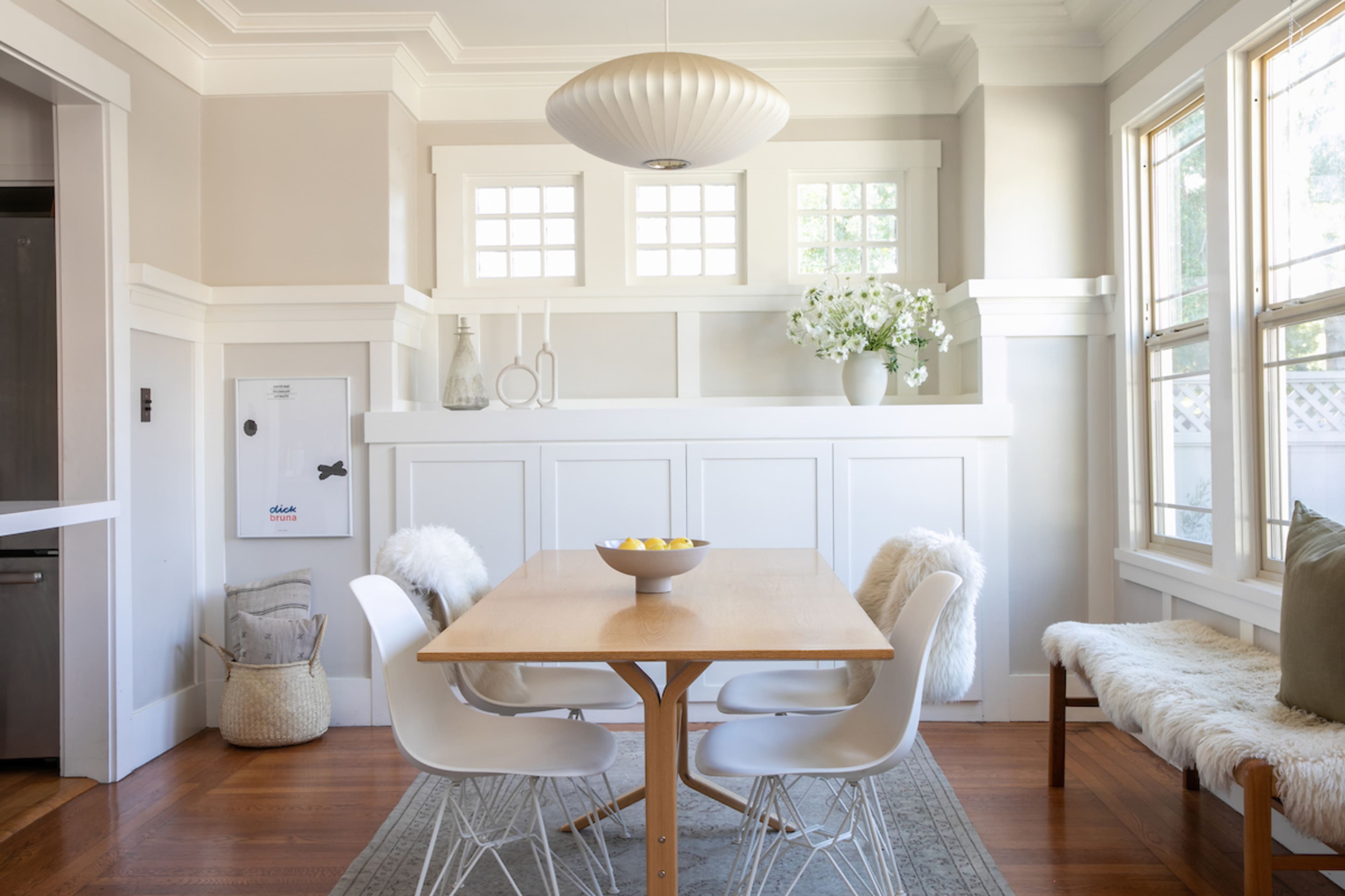 A bright dining area features a wooden table surrounded by four white chairs, with a modern pendant light overhead and a decorative arrangement of flowers and fruit.