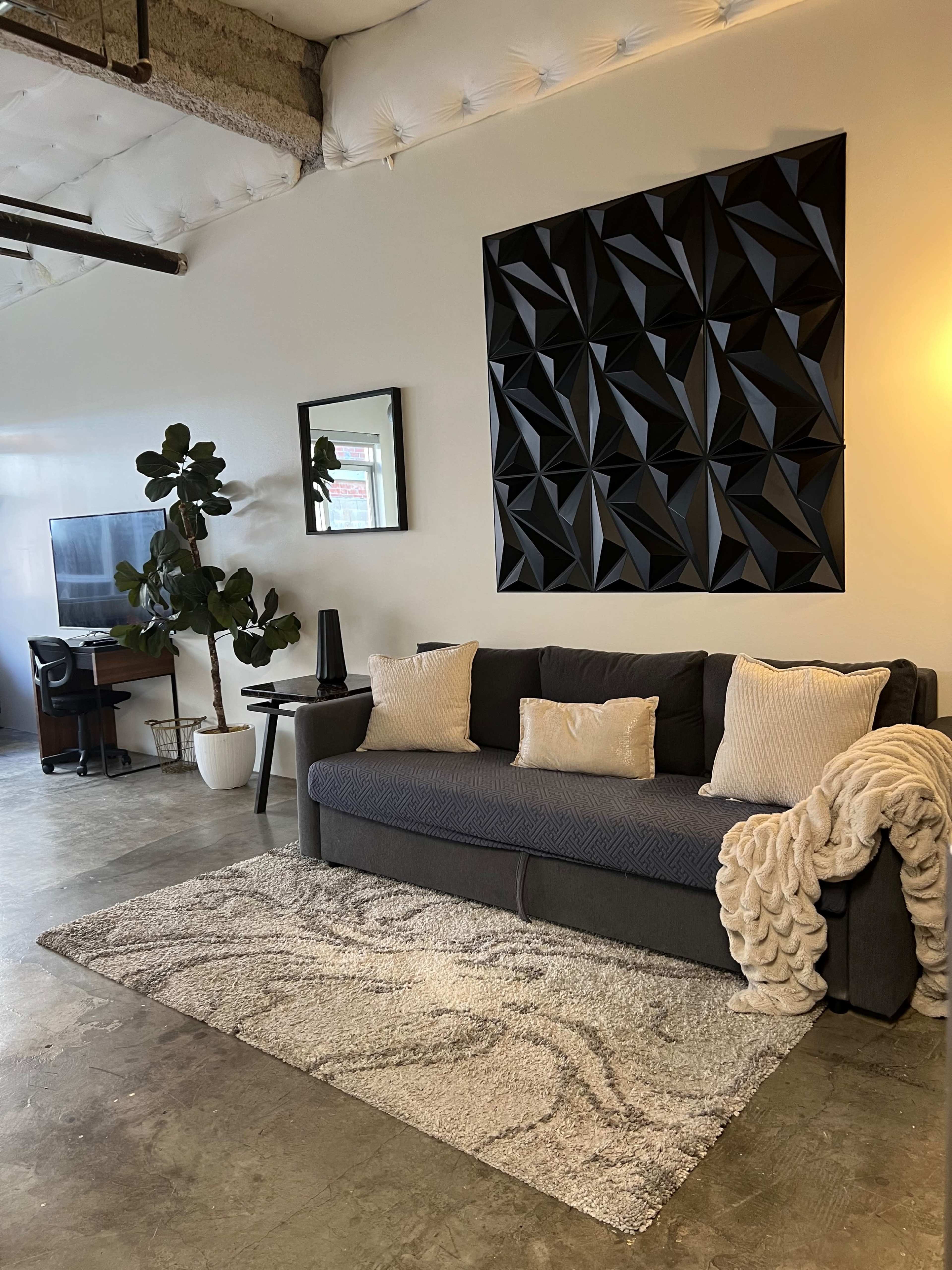 The image depicts a modern living space featuring a dark gray sofa, a geometric black wall art piece, a desk with a computer, and plants, all arranged on a textured rug with concrete flooring.